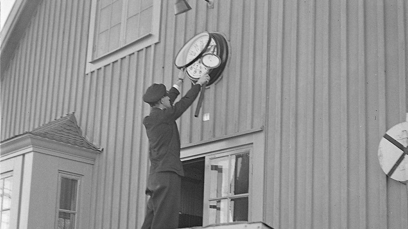 Balck and white picture of a man standing on a roof adjusting a station clock.