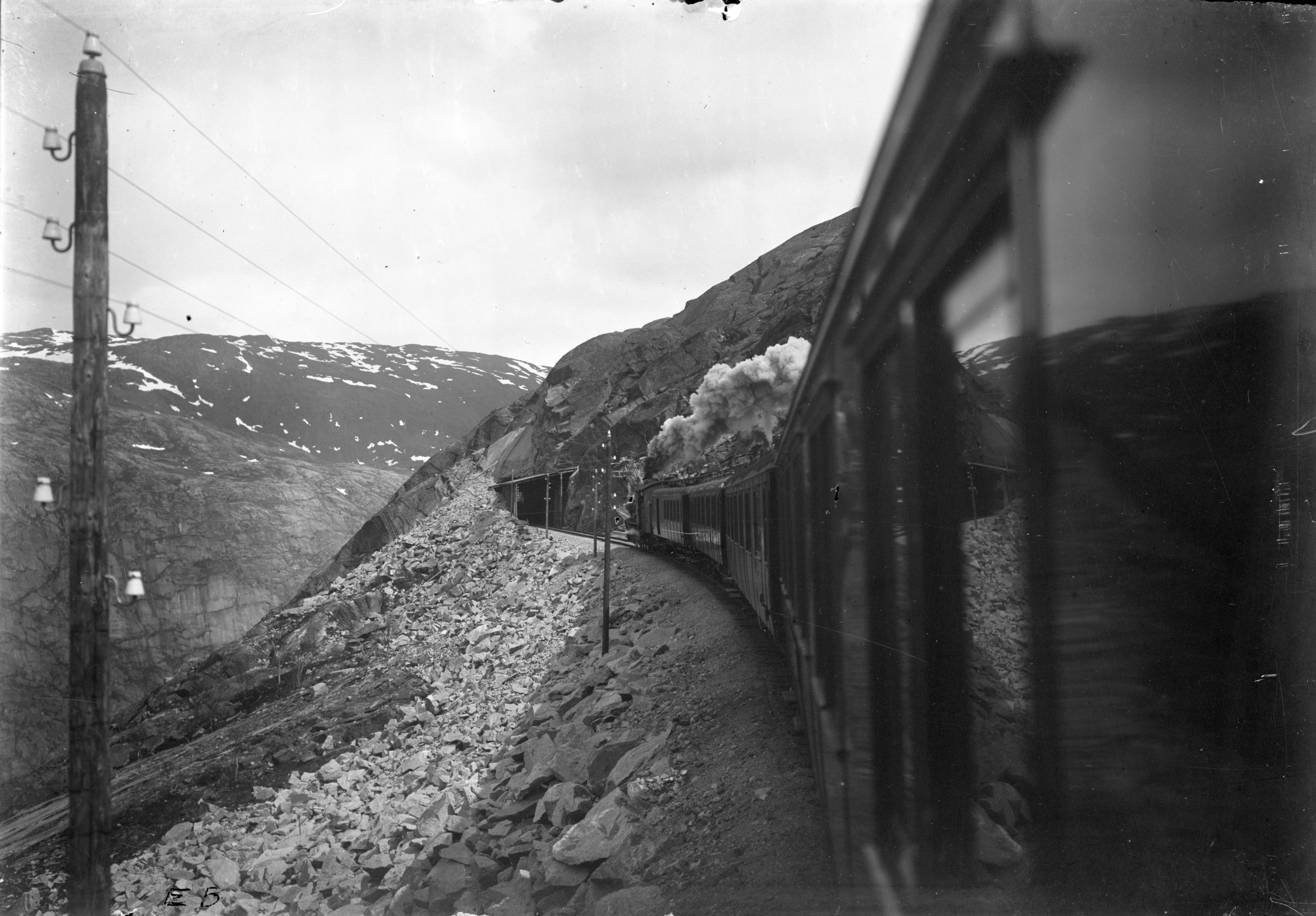 Ett ånglok med vagnar på väg in i en tunnel, det omgivande landskapet visar fläckvis snötäckta fjäll.