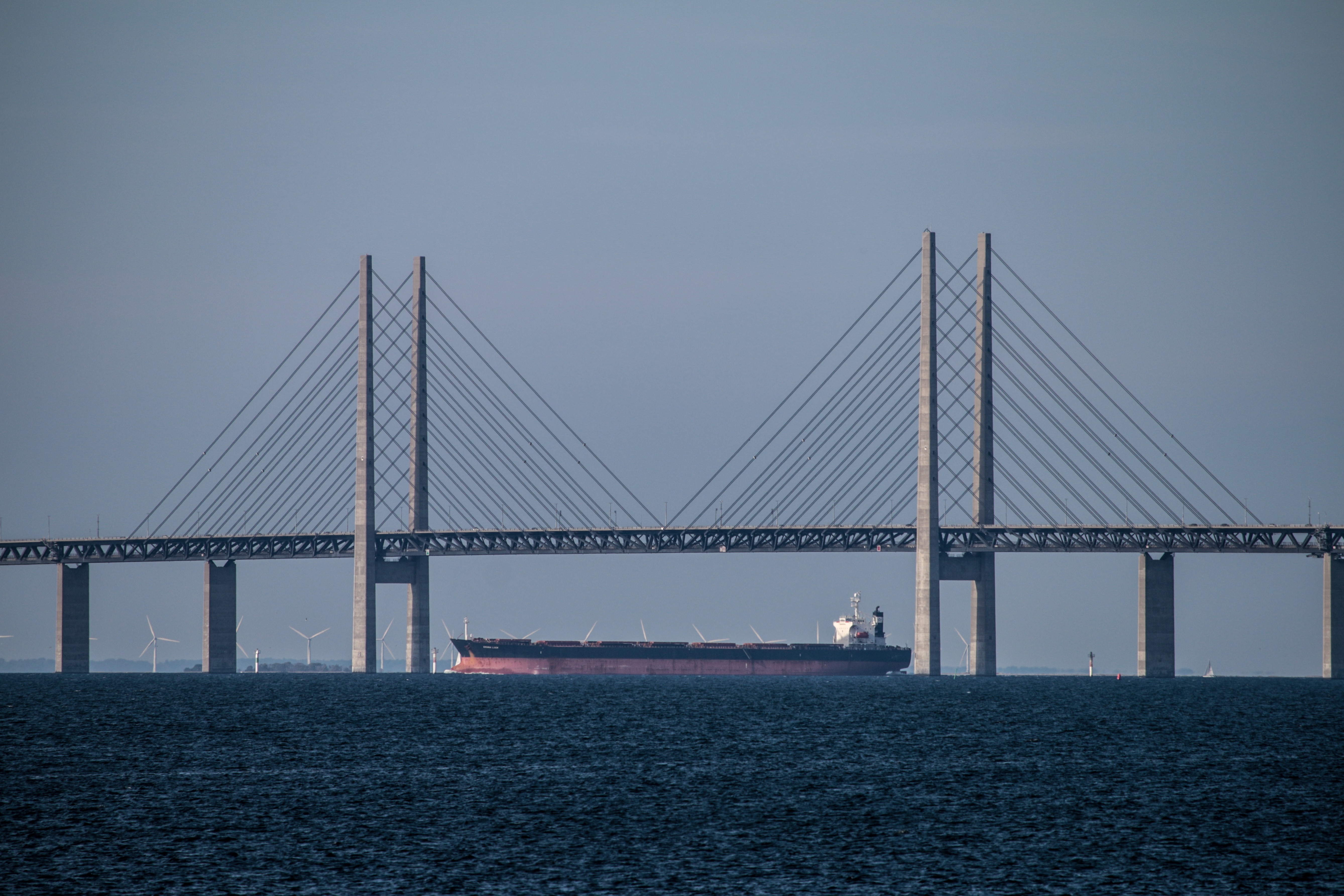 A red-and-black tanker sails under the middle section of the Öresund Bridge in dark water with wind turbines strewn in the background
