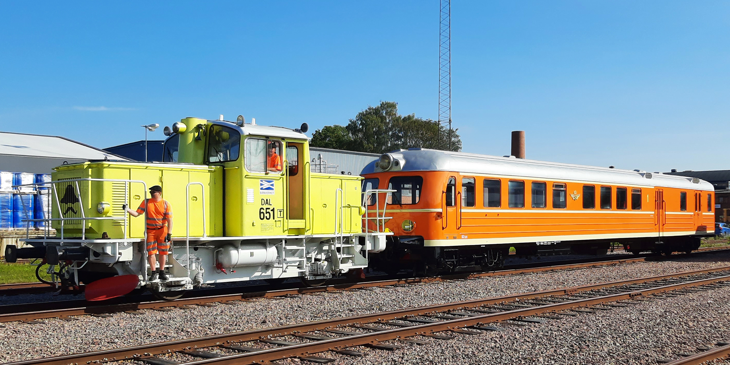 Limegult lok med orange rälsbuss kopplad. En man i förarhytten, en man står längst fram på loket.