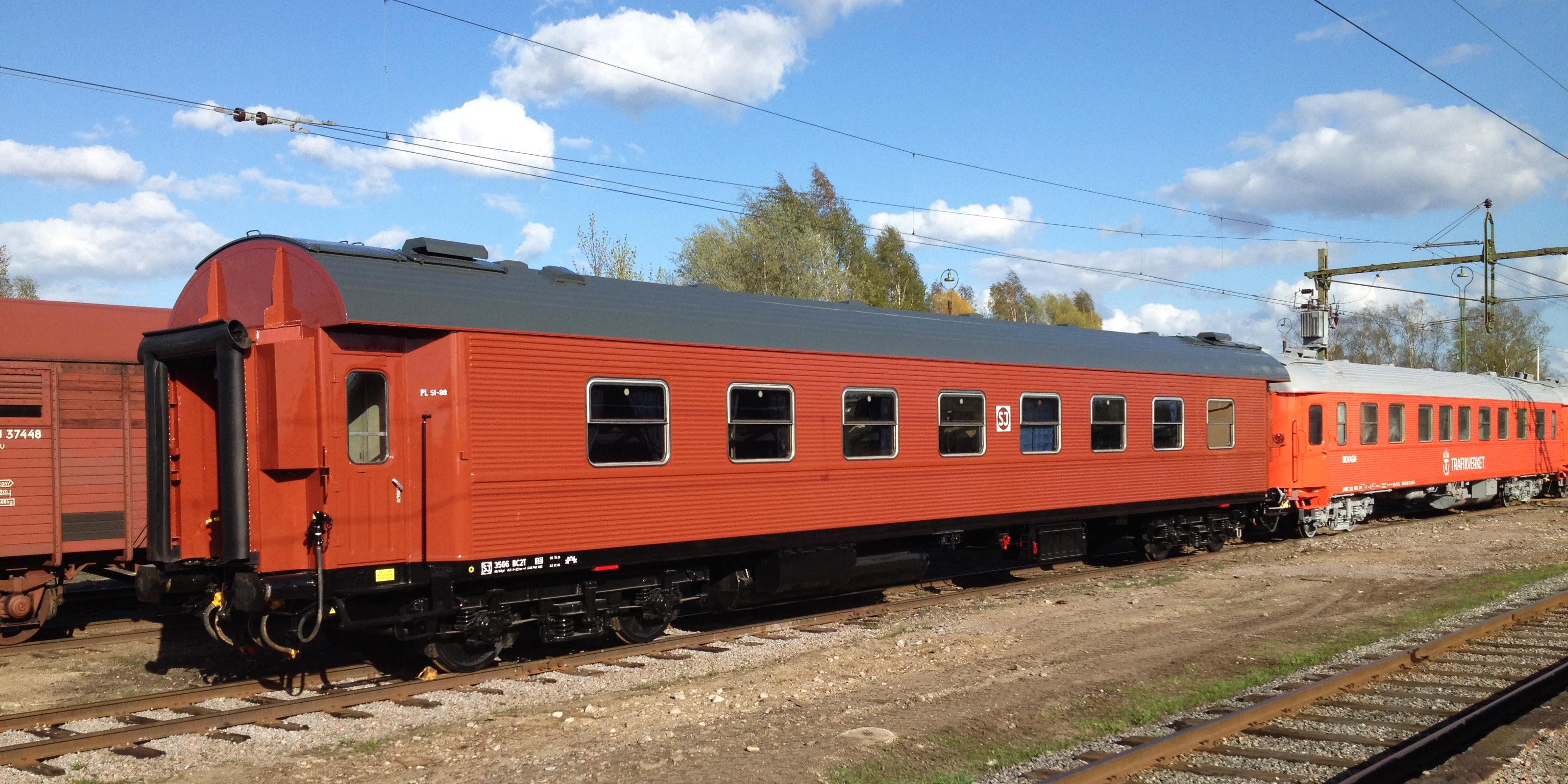 På spåret en röd SJ-tågvagn med en ljusare orange vagn därefter. På parallellspåret bakom skymtar ett godståg.