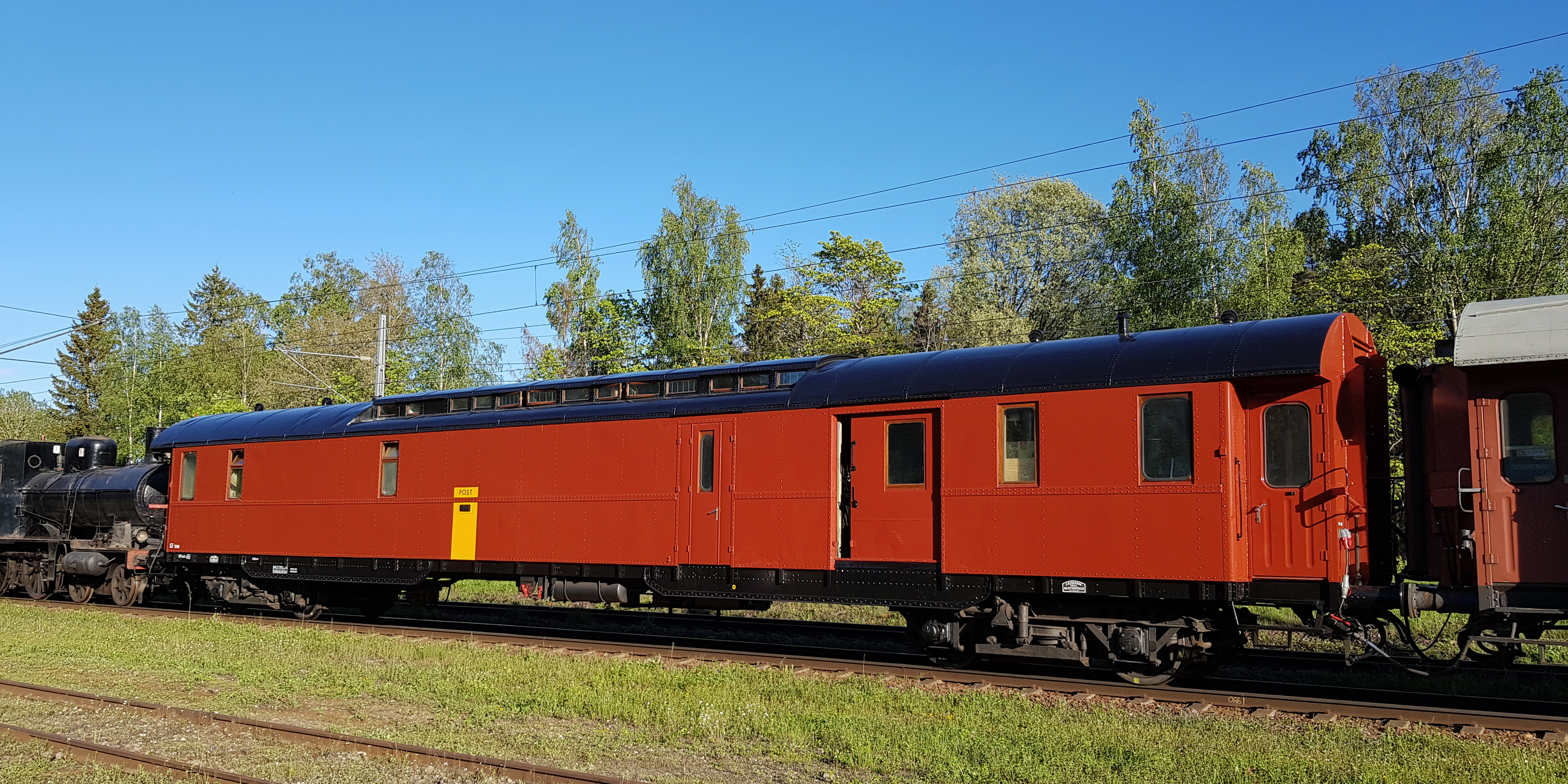 En röd postvagn med få fönster som dras av ett svart ånglok.