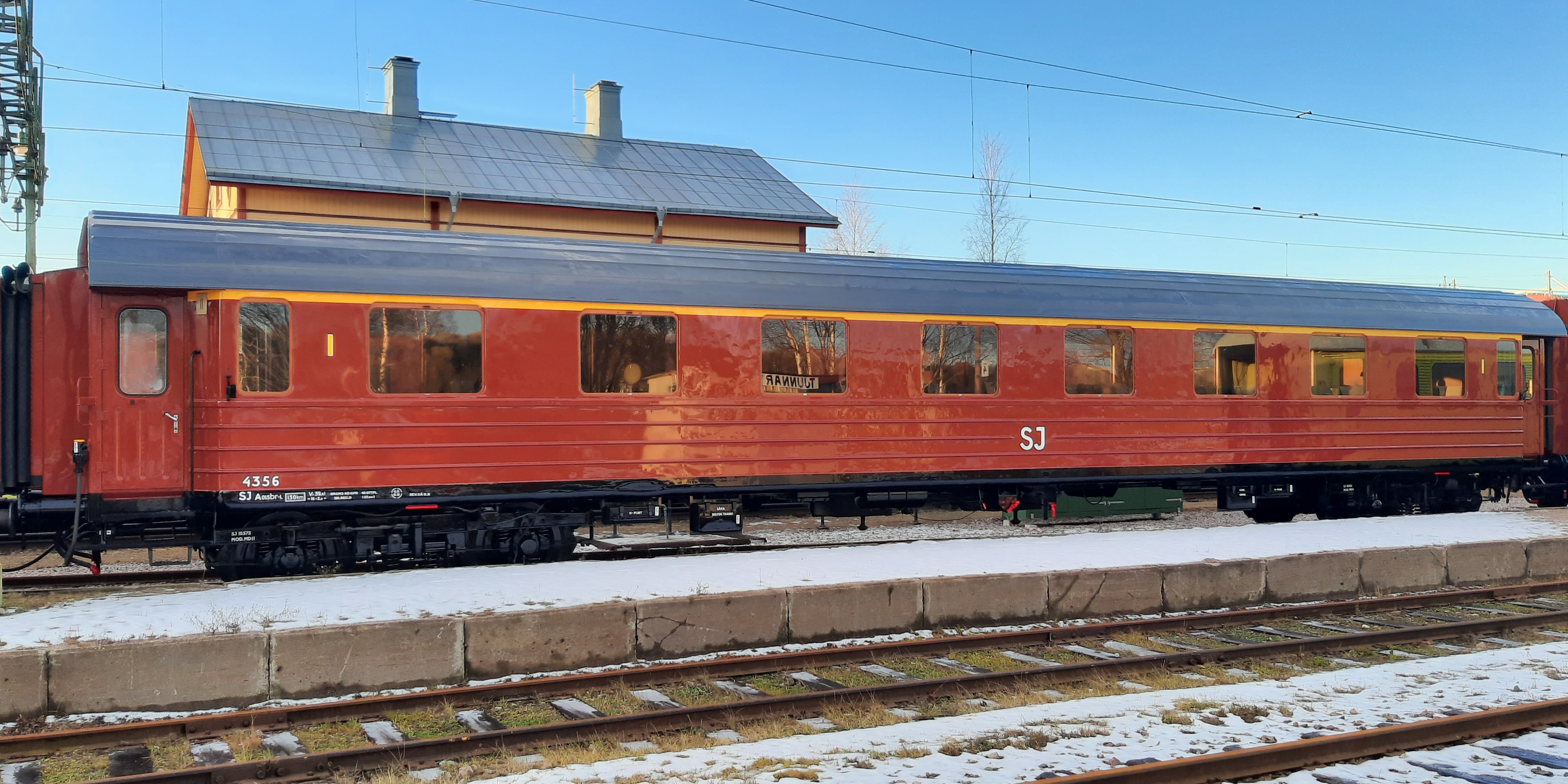 Roströd personvagn med gult streck ovanför fönstren kopplad mellan andra vagnar på spårområde.