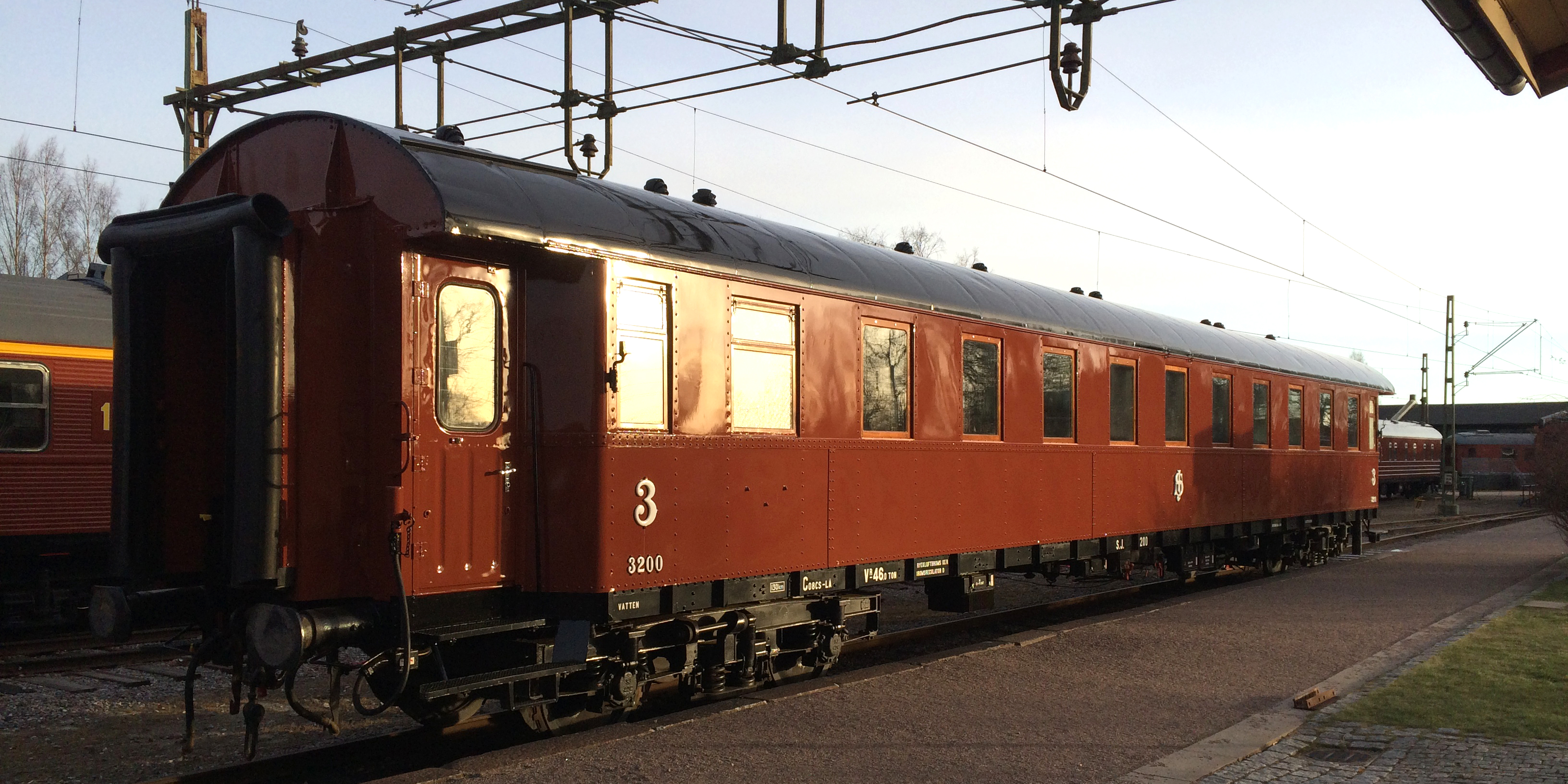 Roströd personvagn med gult streck ovanför fönstren placerad på stationsområde.