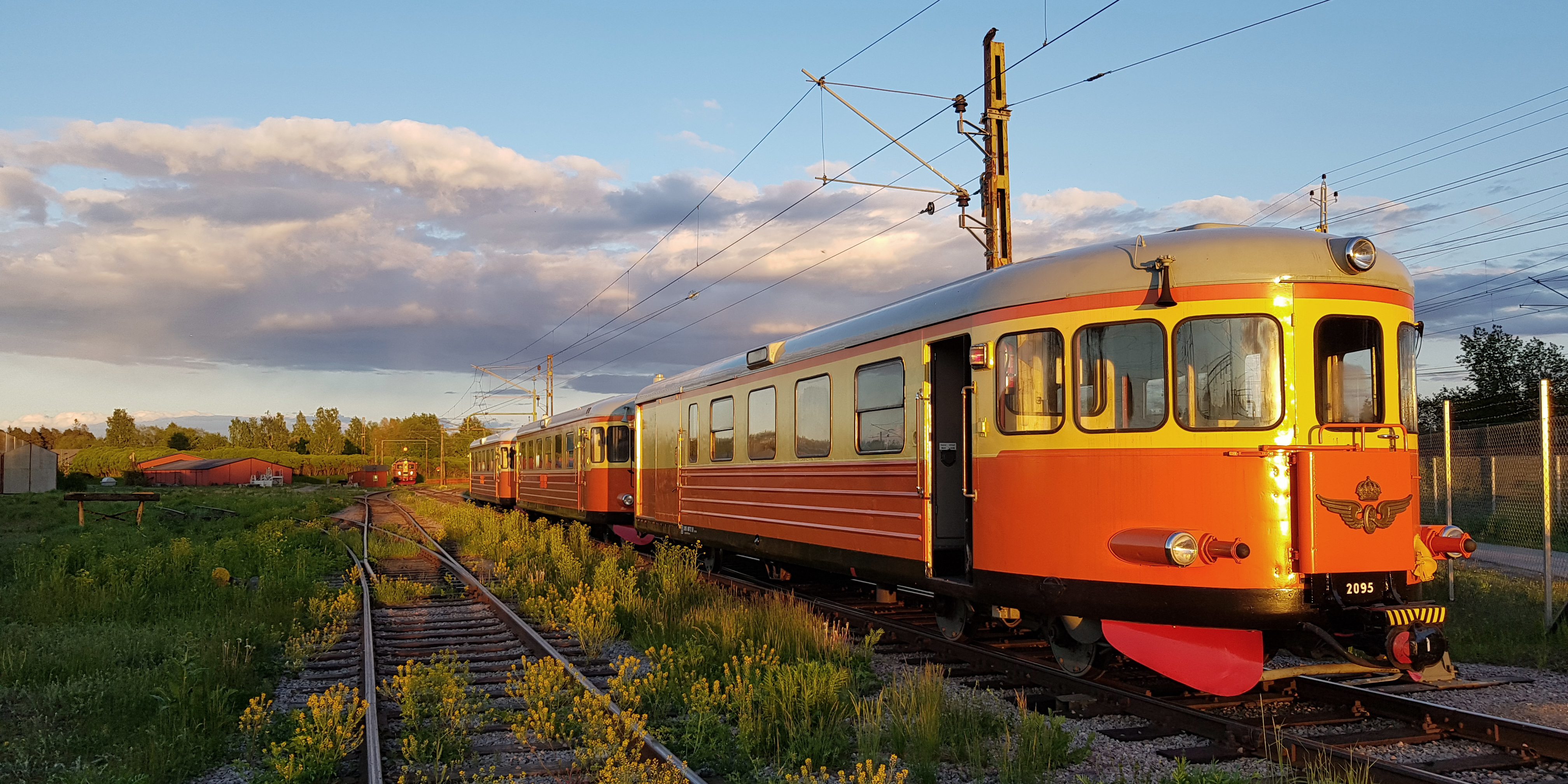 Orange-gul rälsbuss med flera vagnar på rad på banvall i kvällsljus. 