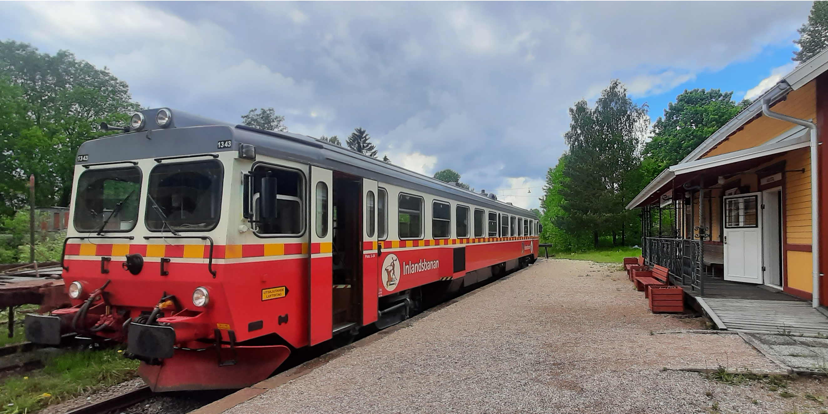 Rödvitt persontåg parkerat med öppna dörrar framför mindre station.
