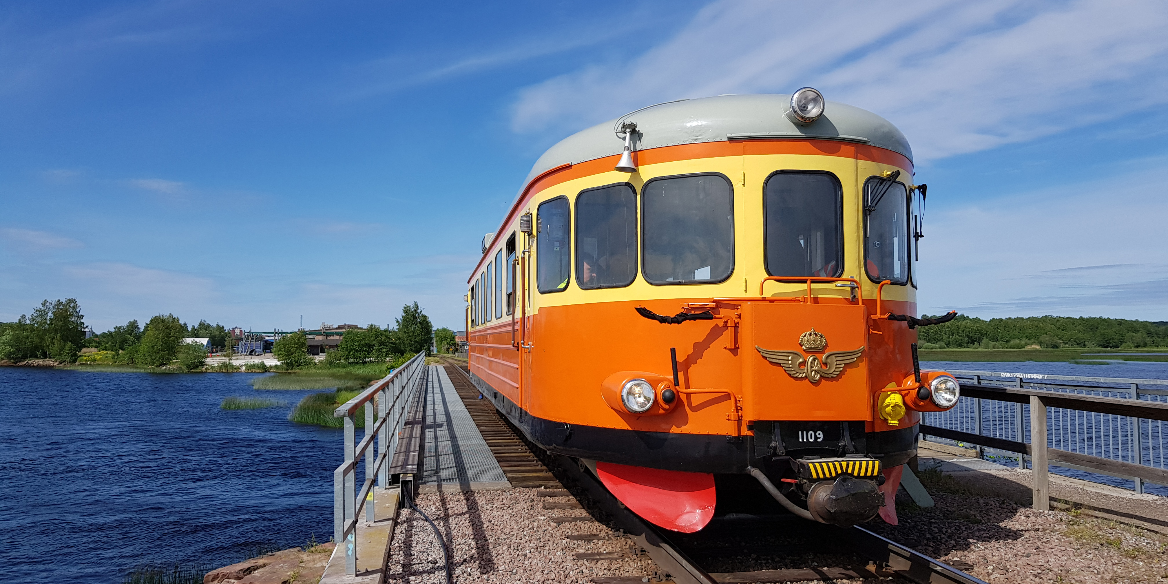 Orange-gul rälsbuss på bro över vatten en sommardag.