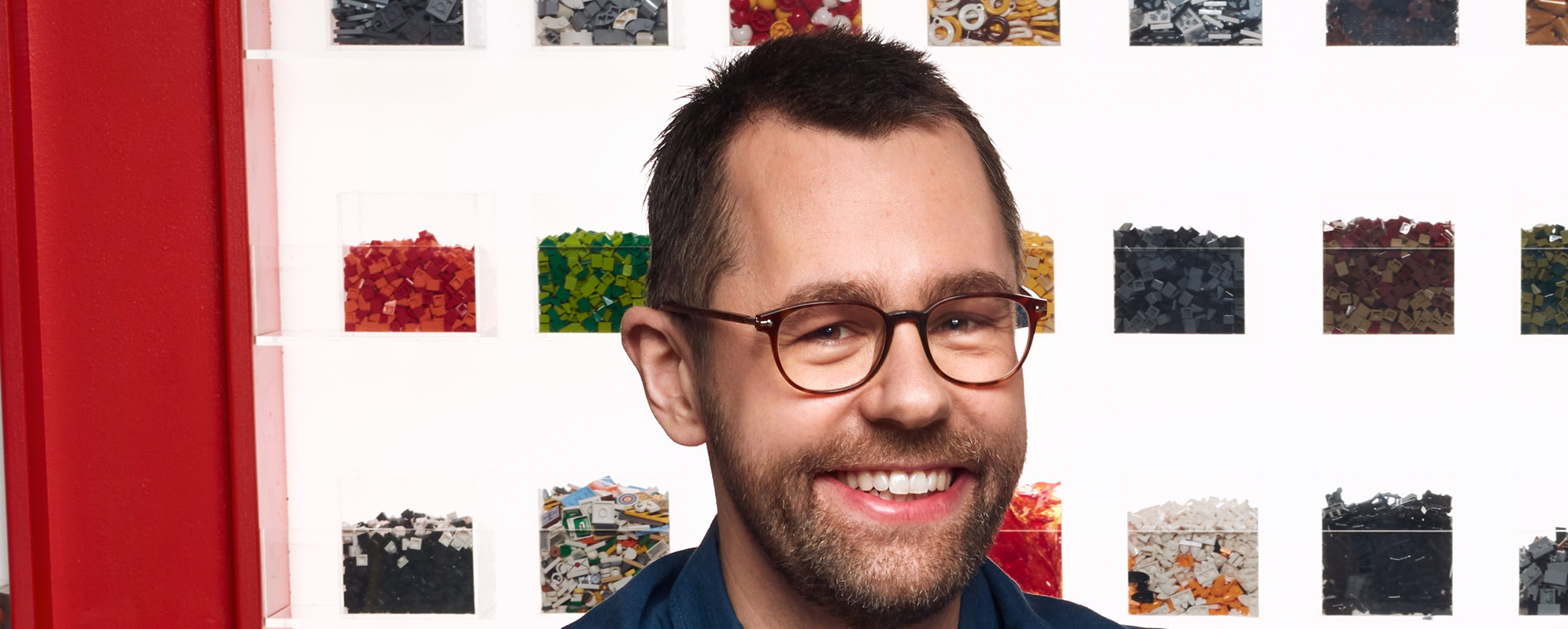 En man i porträtt. Kortklippt hår och mörkblå pikétröja. I bakgrunden syns Lego sorterat i lådor.