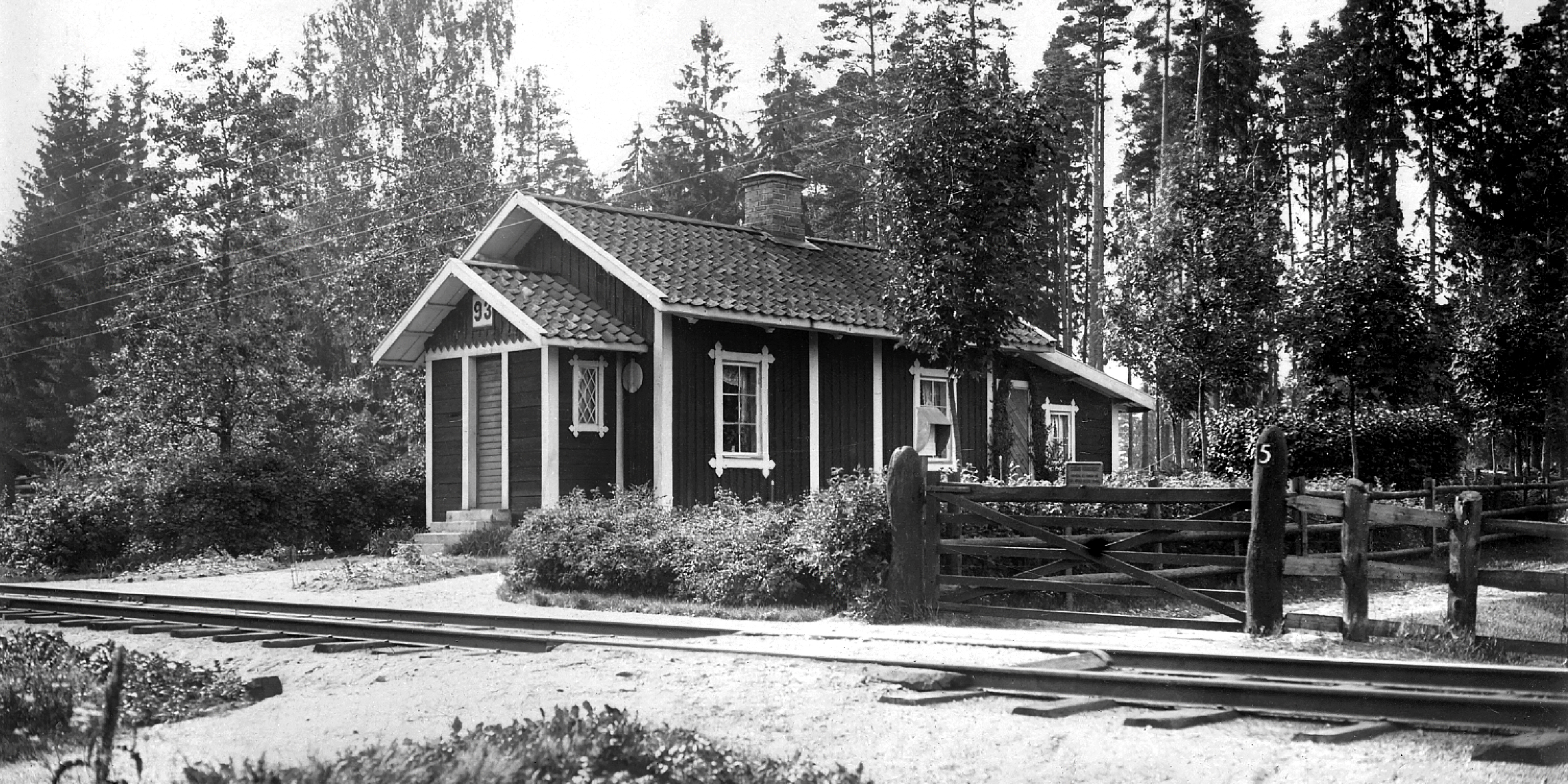 Trähus med ljusa knutar framför järnvägsspår. Vägövergång med trägrind i förgrunden.
