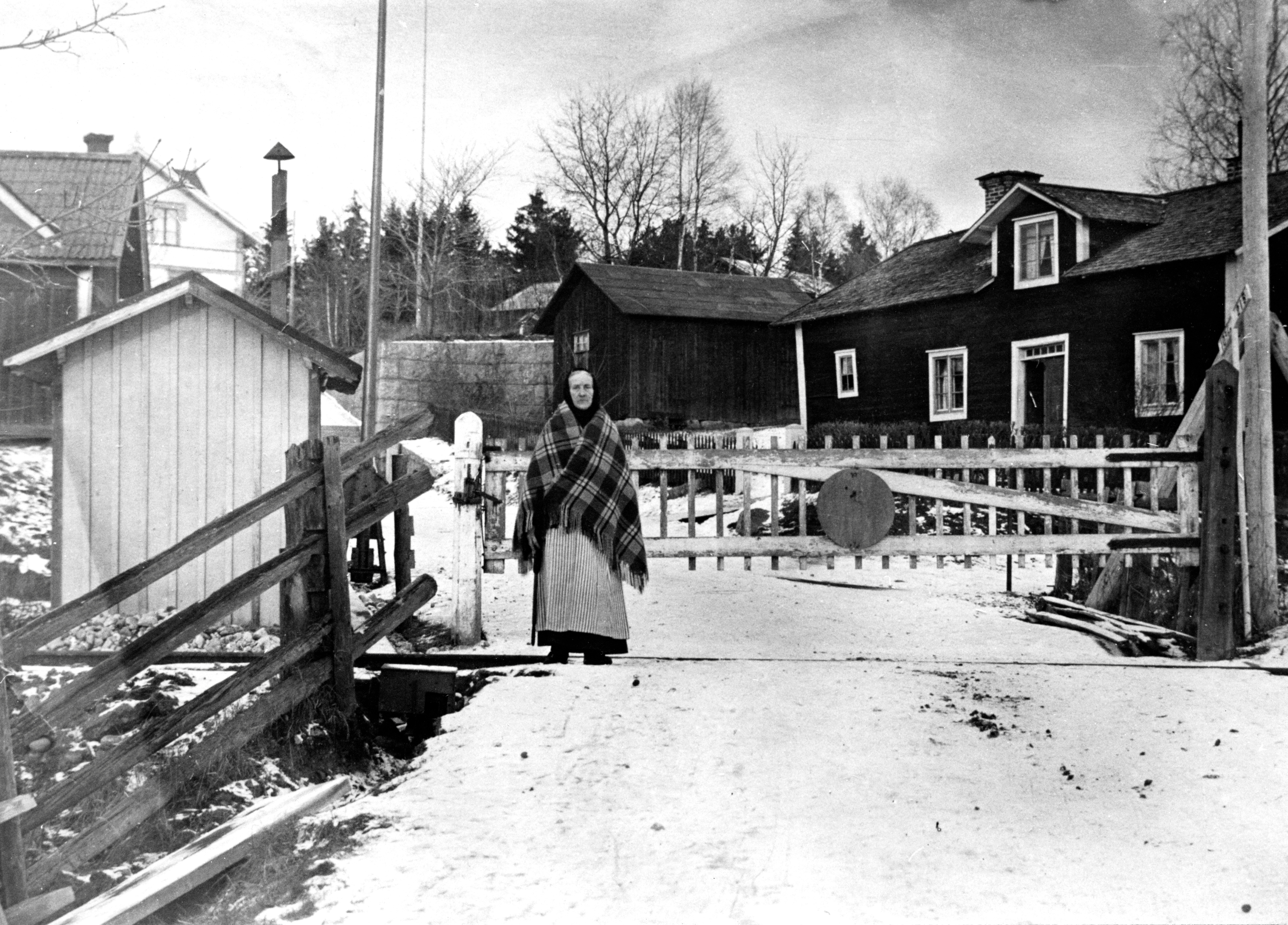 Kvinna stående på väg framför järnvägsgrind med schal runt axlarna i vinterlandskap. Trähus i bakgrunden.