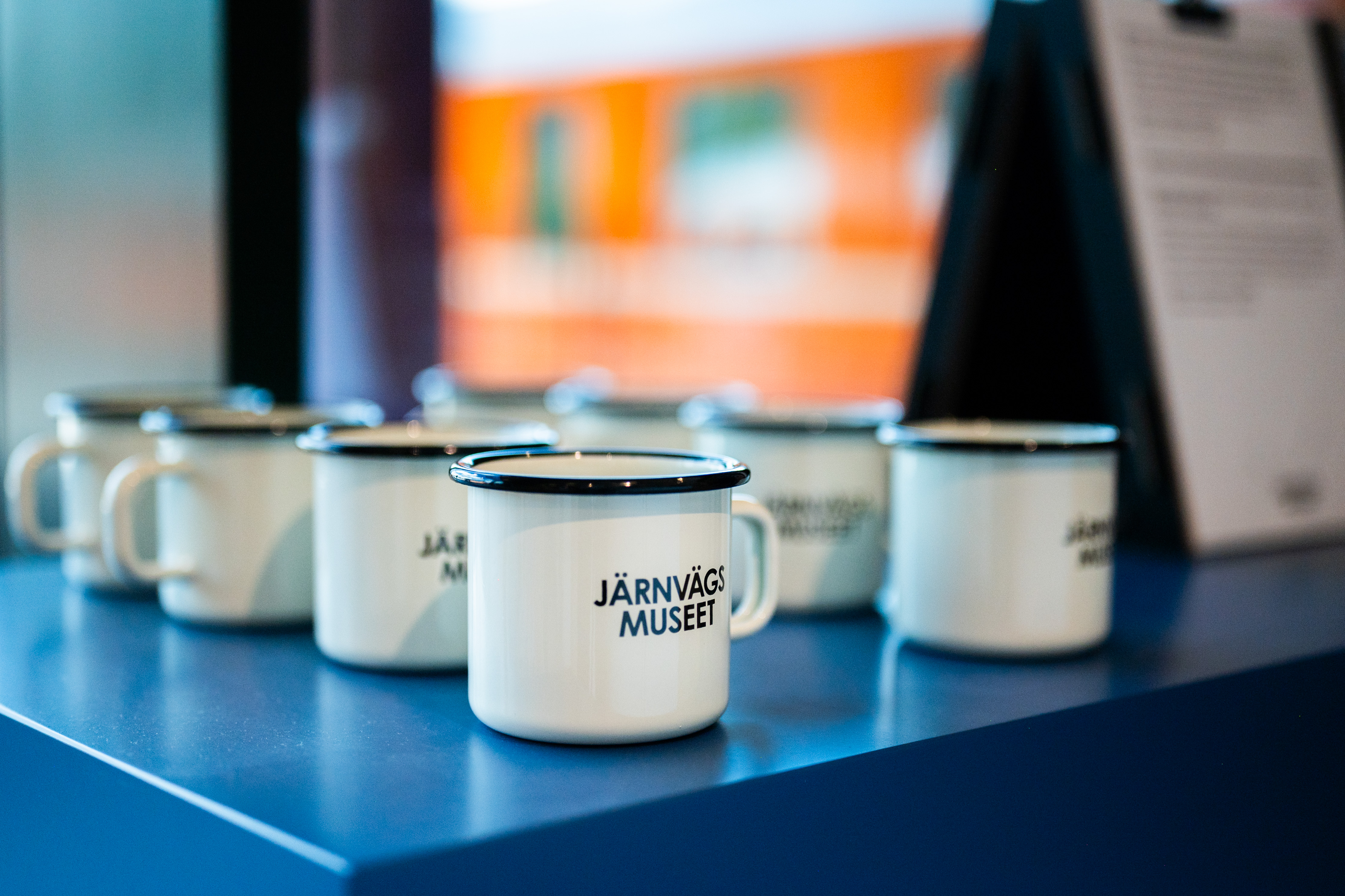Åtta svartvita plåtmuggar med texten Järnvägsmuseet, står uppradade på en blå hylla.