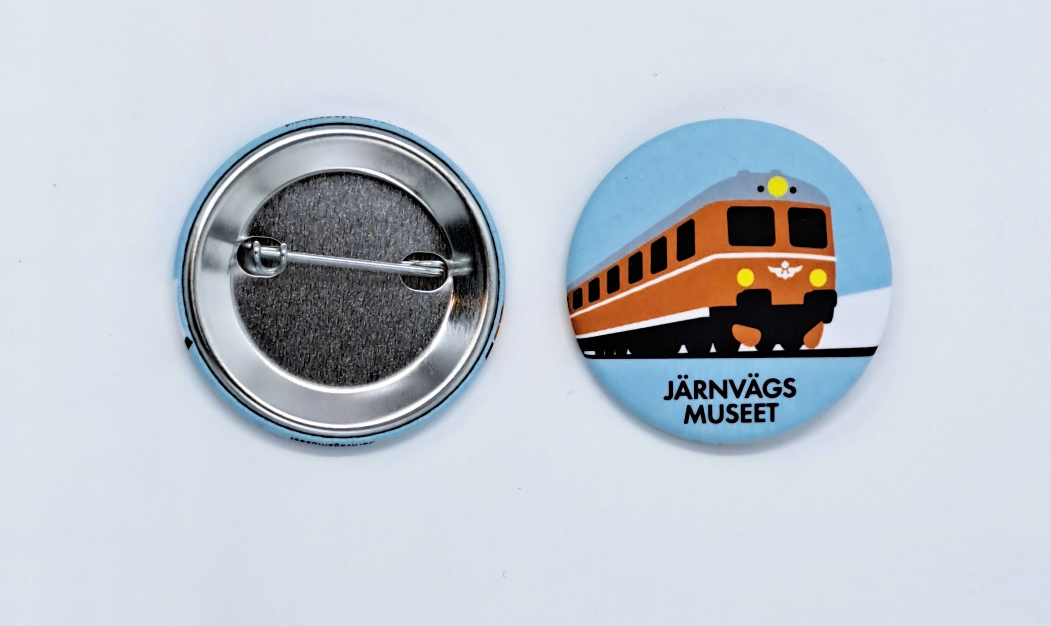 En rund pin med ljusblå bakgrund och en röd motorvagn som motiv. Nedtill står det "Järnvägsmuseet".