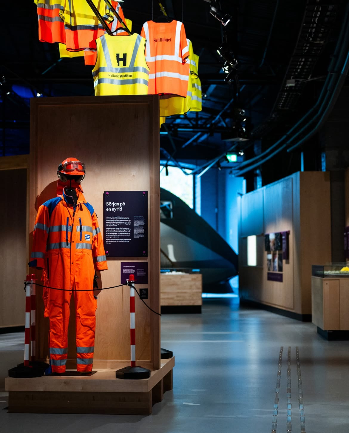 En museiutställning som visar en orange skyddsoverall med reflexband, uppställd bakom avspärrningar. Bakom montern hänger flera reflexvästar i olika färger och med tryck från exempelvis "Hallandstrafiken" och "Sällskapet". Informationsskylt med texten "Början på en ny tid" är monterad bredvid overallen. Utställningen är upplyst med spotlights i en mörk lokal med träinredning och ett modernt tåg syns i bakgrunden.