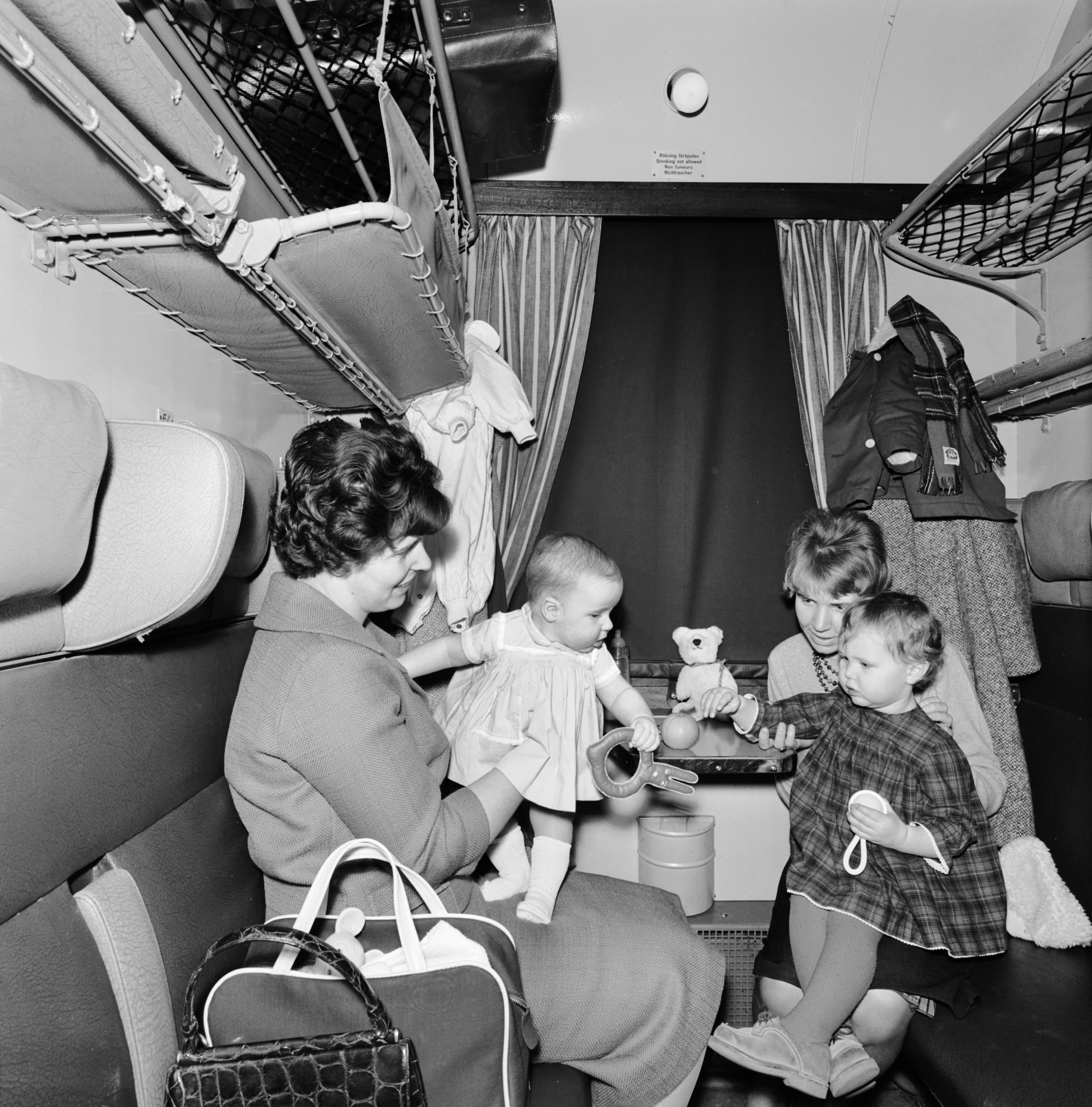 Två kvinnor sitter mittemot varandra i en tågkupé med två små barn. En kvinna håller en bebis, medan den andra hjälper ett äldre barn vid bordet. En nallebjörn och en nappflaska syns på bordet.