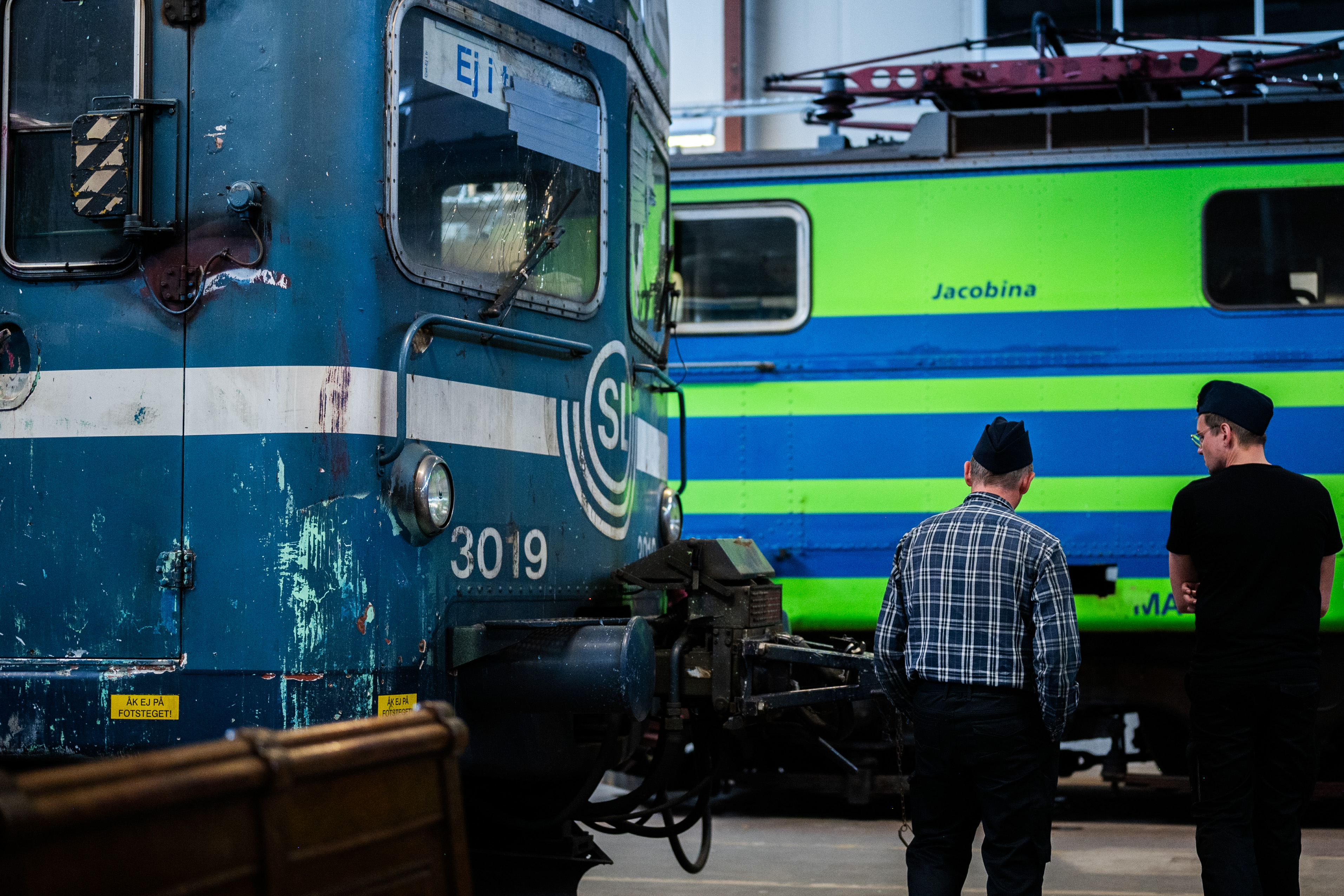A blu train car with the SL logo can be seen in the left corner. To the right two man are standing and watching another train car in the background.