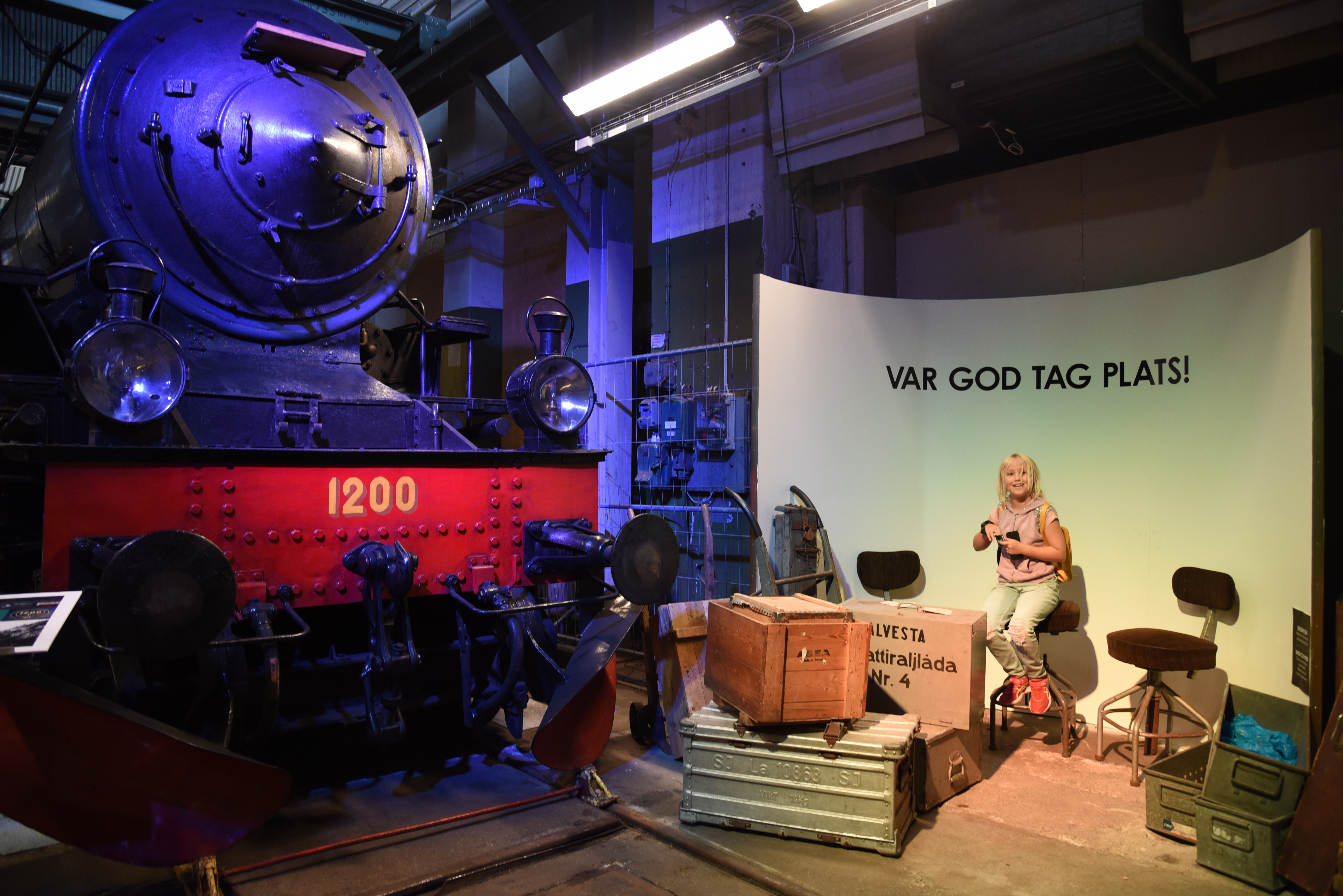 En flicka sitter bredvid ett stort svart lok, bakom henne står det "var god tag plats!"