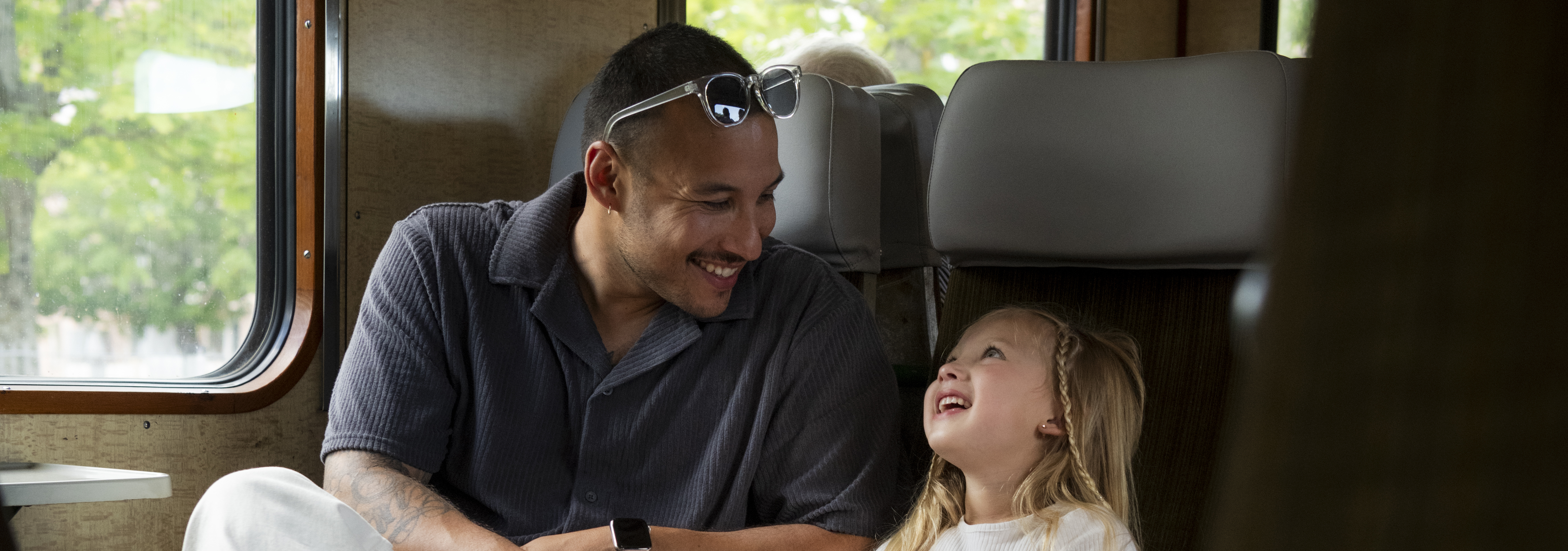 En pappa och dotter sitter i en rälsbussvagn på var sitt säte och kollar leendes på varandra utanför fönstret skymtar grönskande träd.