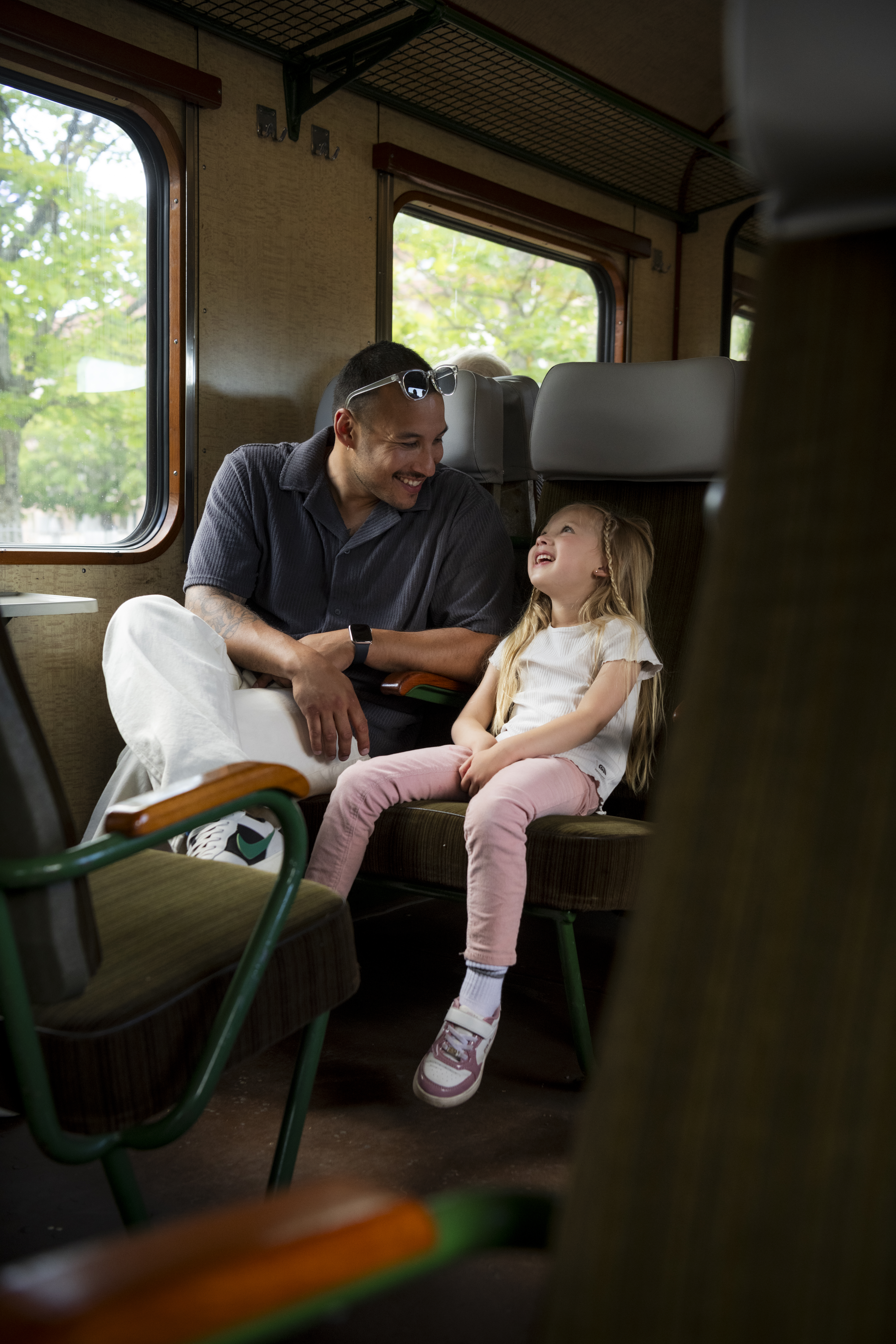 A father and daughter are sitting next to each other in a railcar and smiling. You can see the green trees through the window.