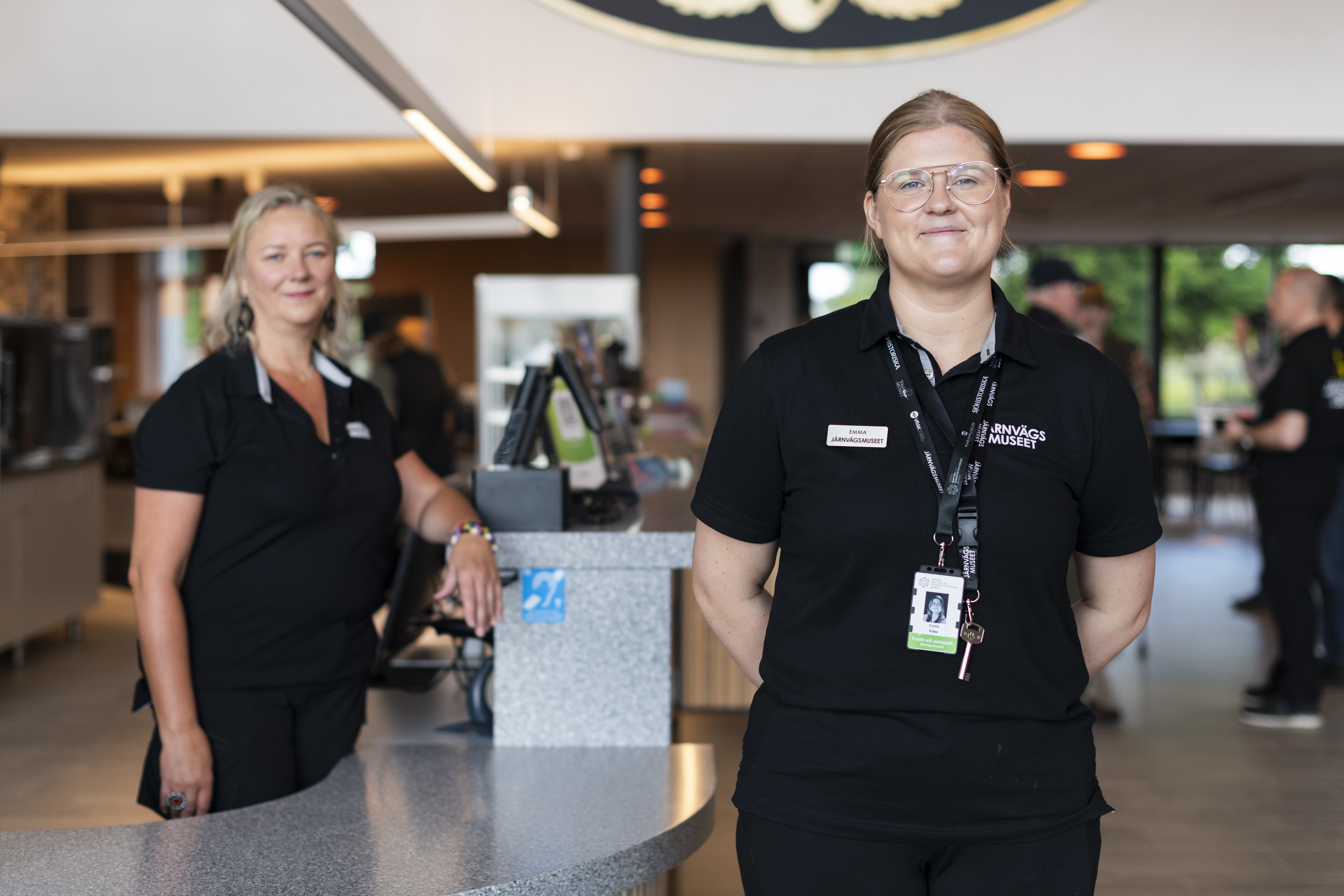En kvinna står och ler in i kameran, hon har en svart tröja med järnvägsmuseets logotyp på