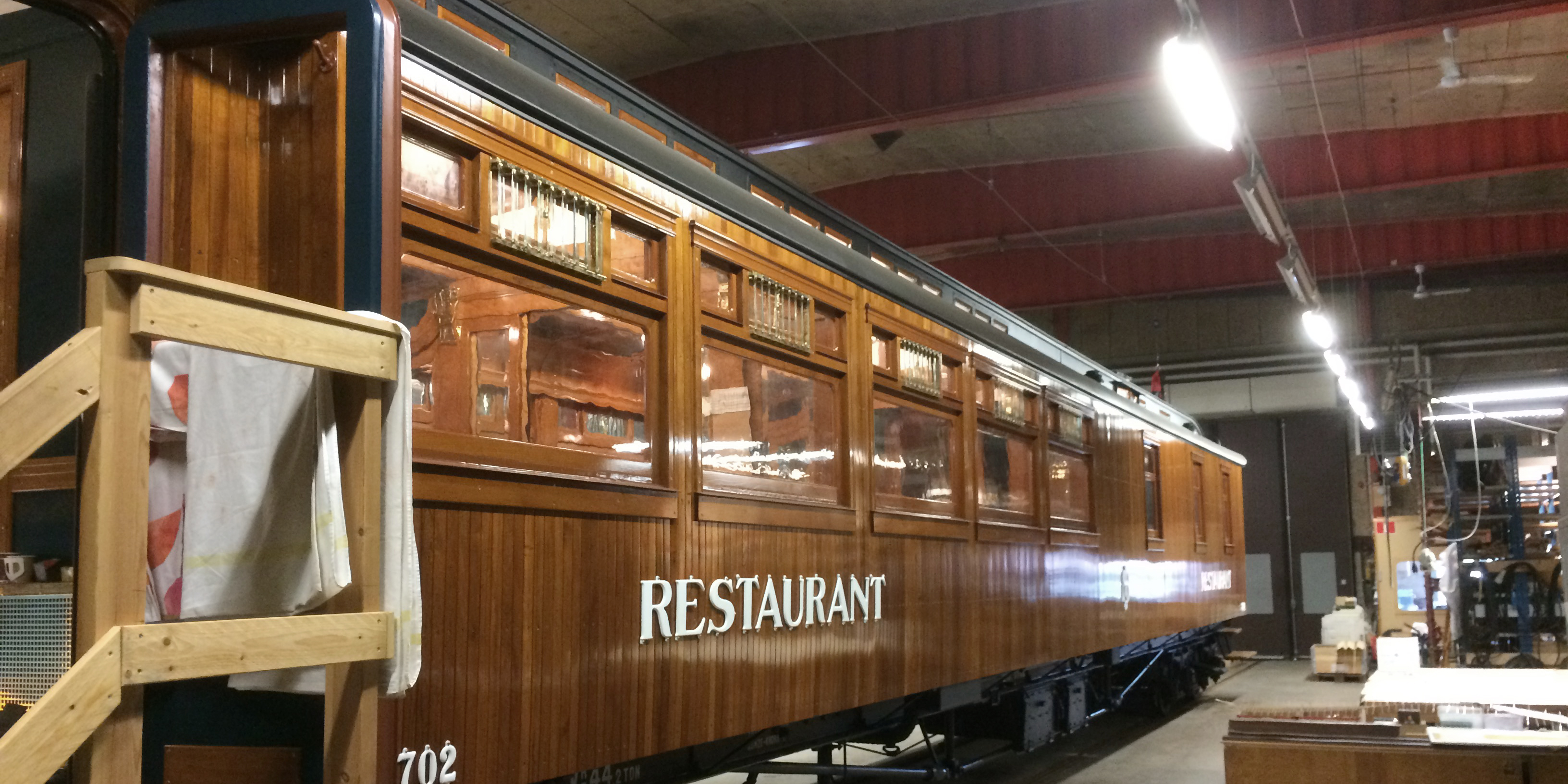 Träpanelad tågvagn i tåghall med ordet Restaurant på sidan. Trapp leder in i vagnen för besökare.