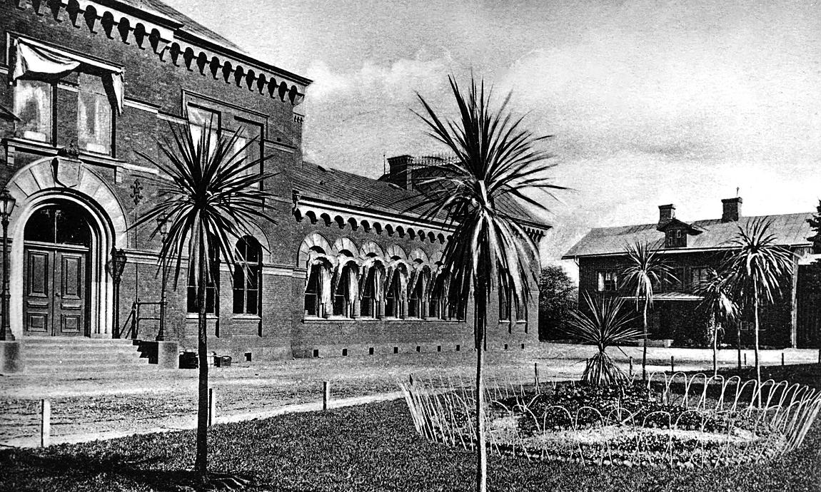 Framsida av stor byggnad med gräsmatta och flertalet palmer som växer.