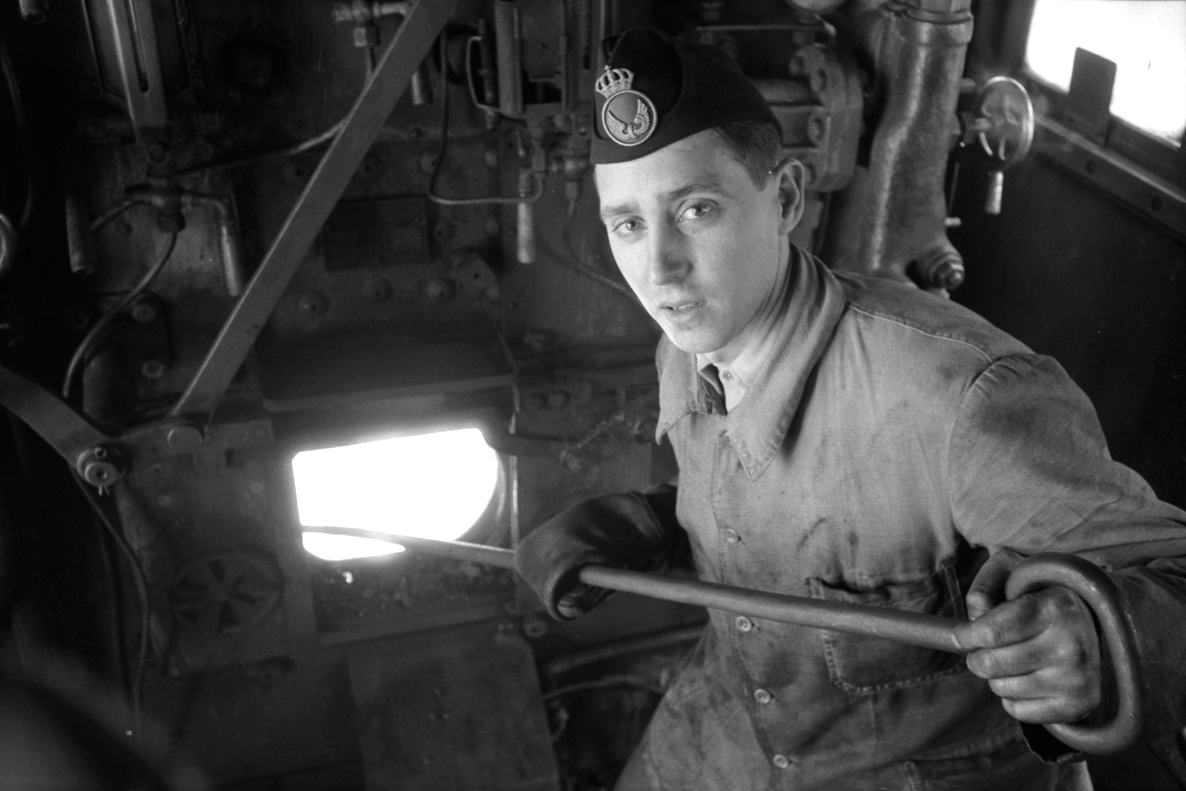 A man by an open fire hatch is used on steam locomotives where fire is burning. He holds iron skewers into the fire. 