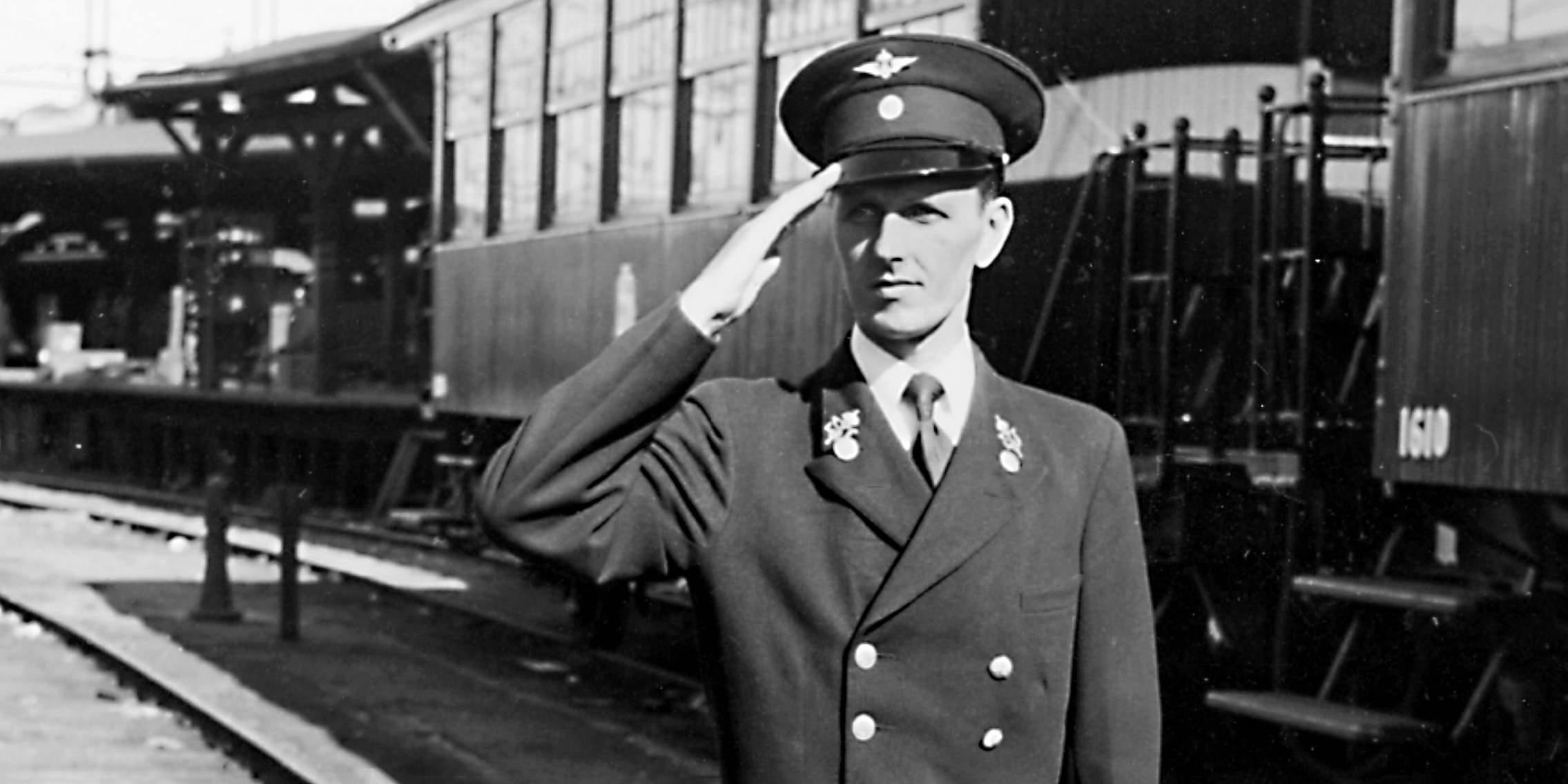 Man in uniform standing at attention in front of trains in the station area.