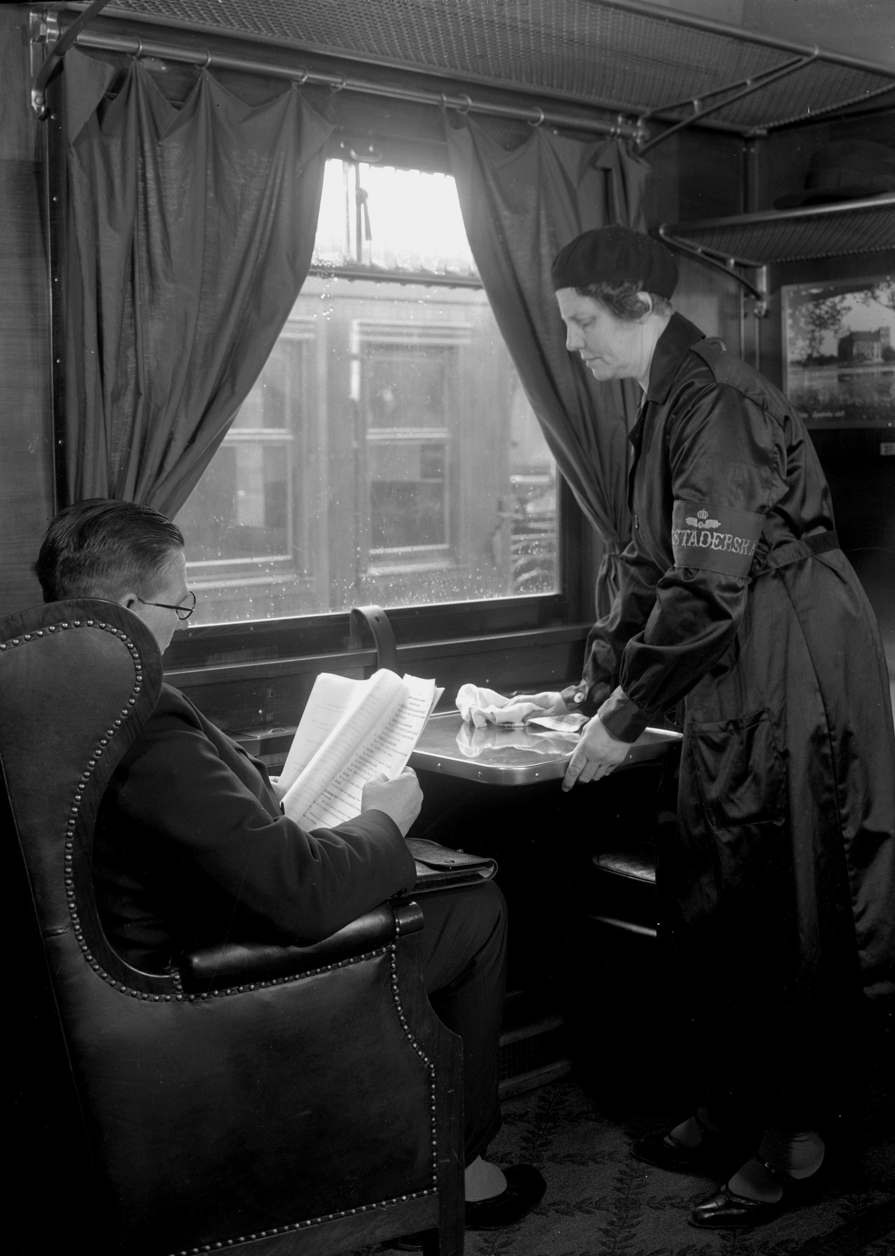 Kvinna torkar av bord framför sittande man i tågkupé.