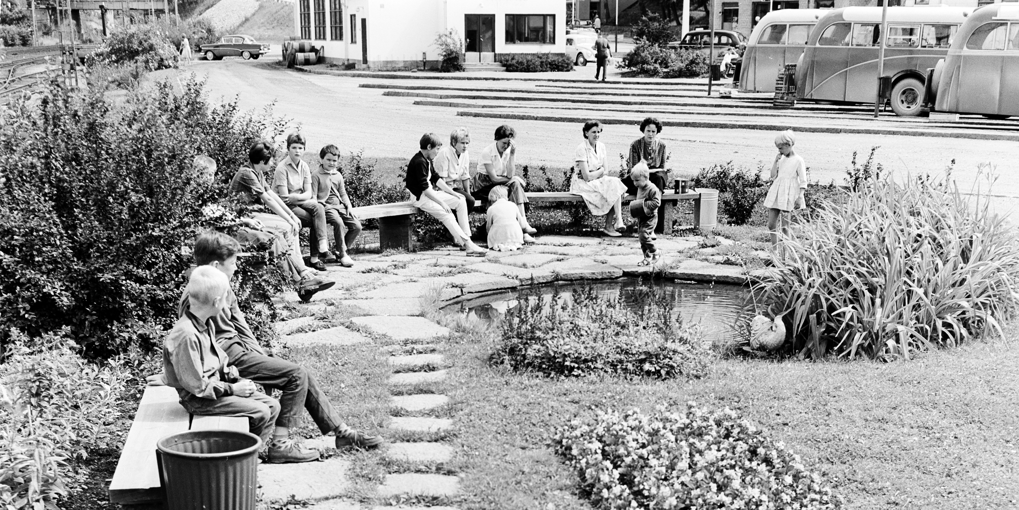 Children and adults sitting on benches in a summery park. Buses in the background.