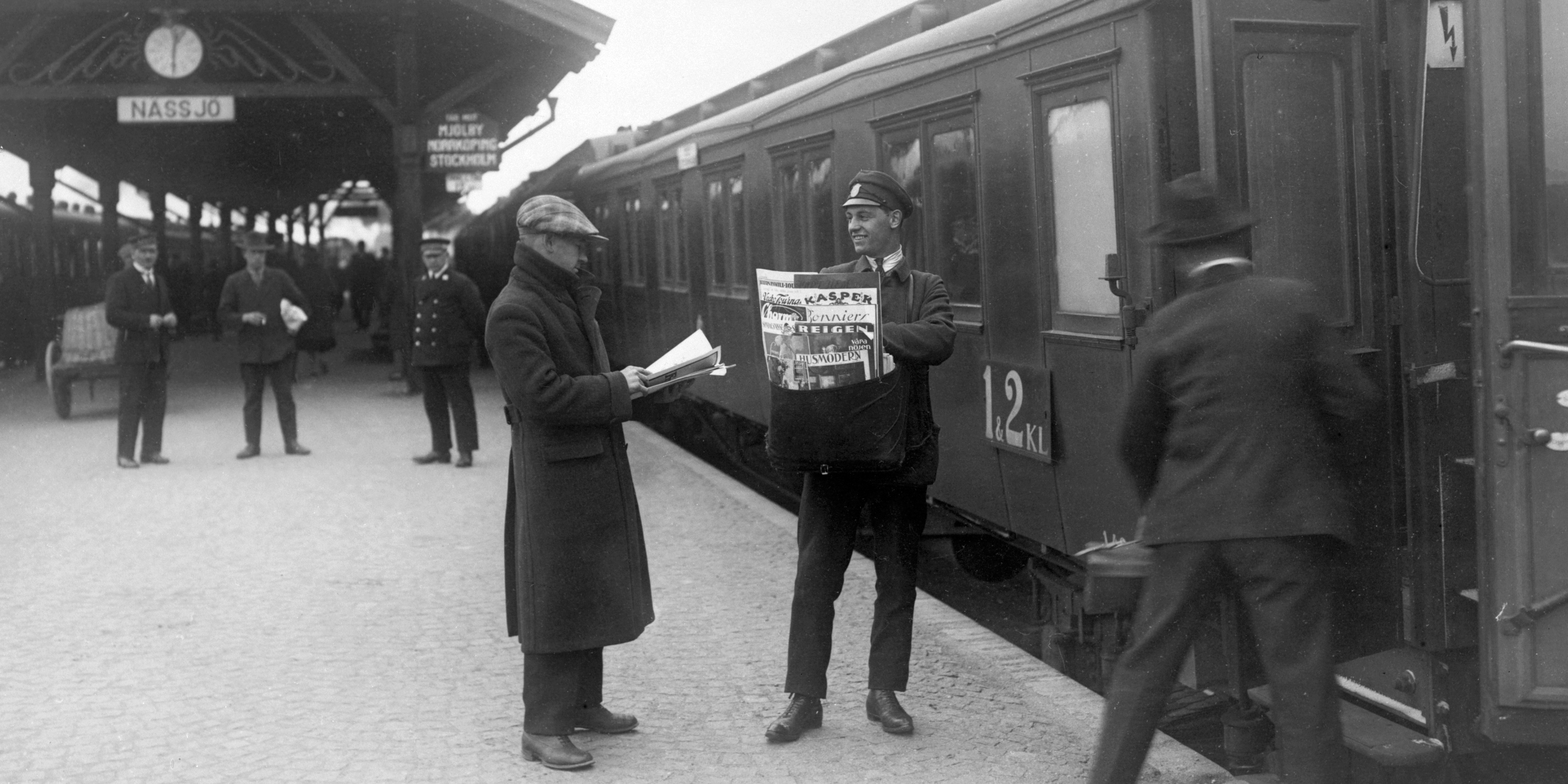 En uniformerad man säljer tidningar på en perrong vid ett avgående tåg och mitt emot honom en kund i kappa och keps läser en dagstidning.