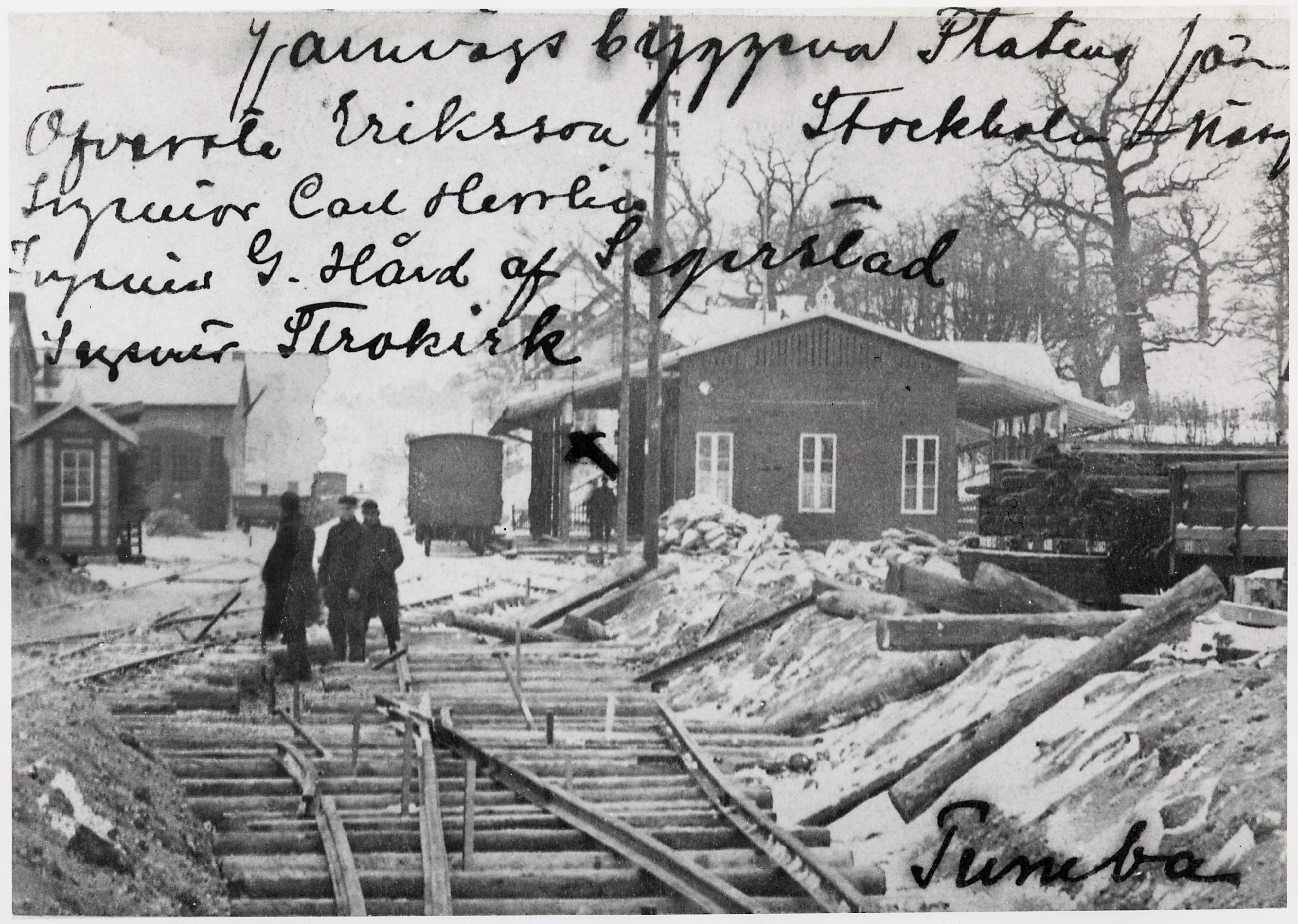 Navvies pause a trail construction in snow with a few houses in the background.