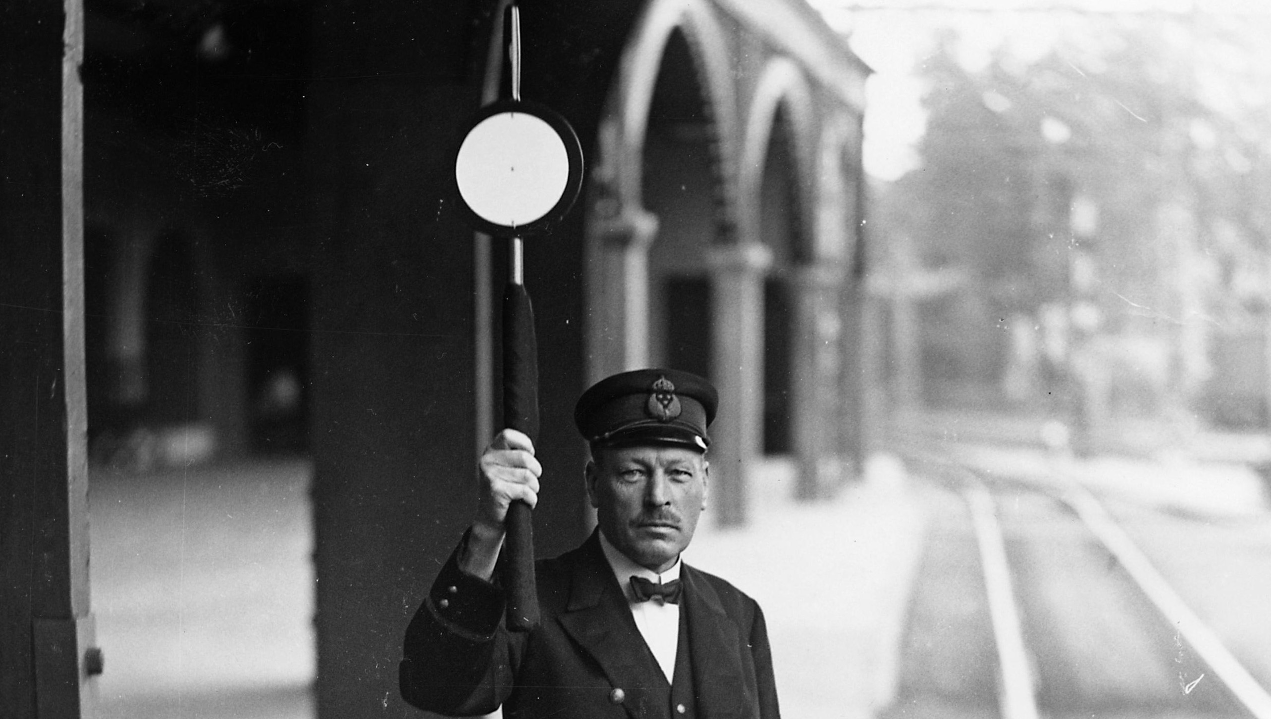 Tågklarerare eller stins står på perrong och håller upp en signalstav vertikalt med höger hand.