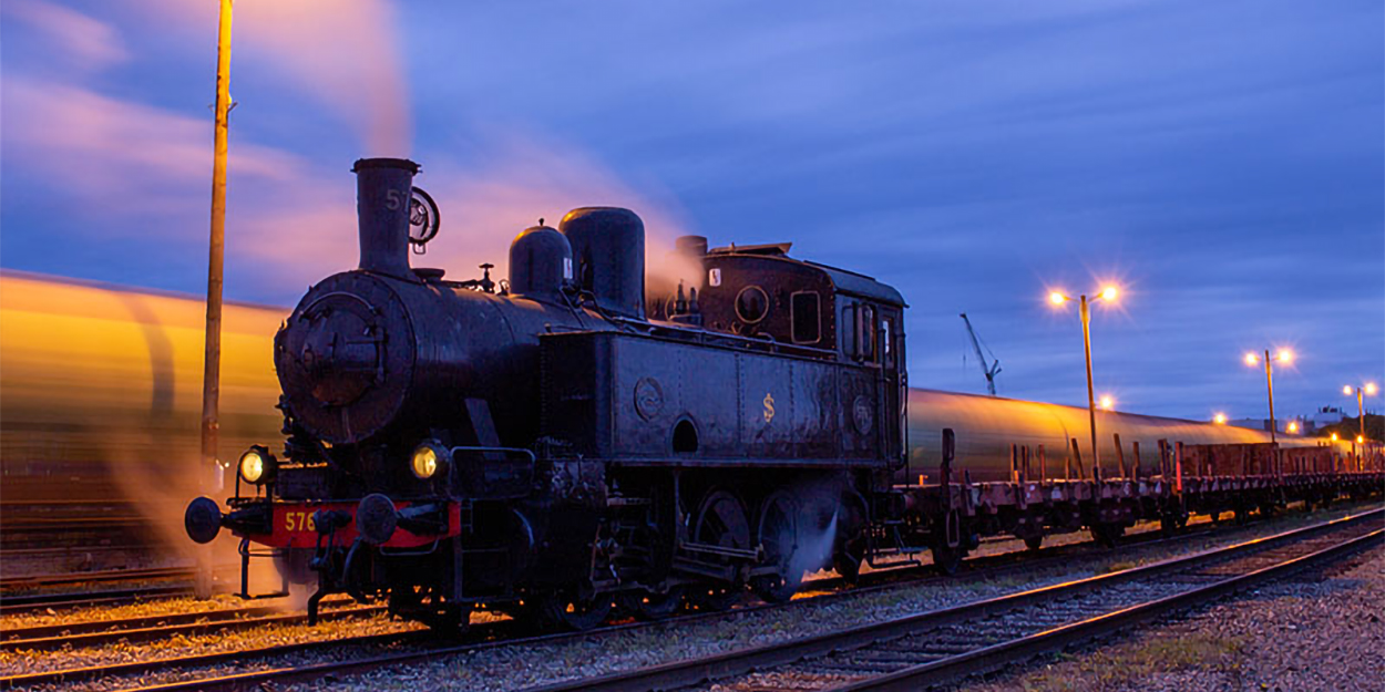 Lok med många godsvagnar i kvällsljus på järnvägsspår. Lång exponering, ljusstrimmor.