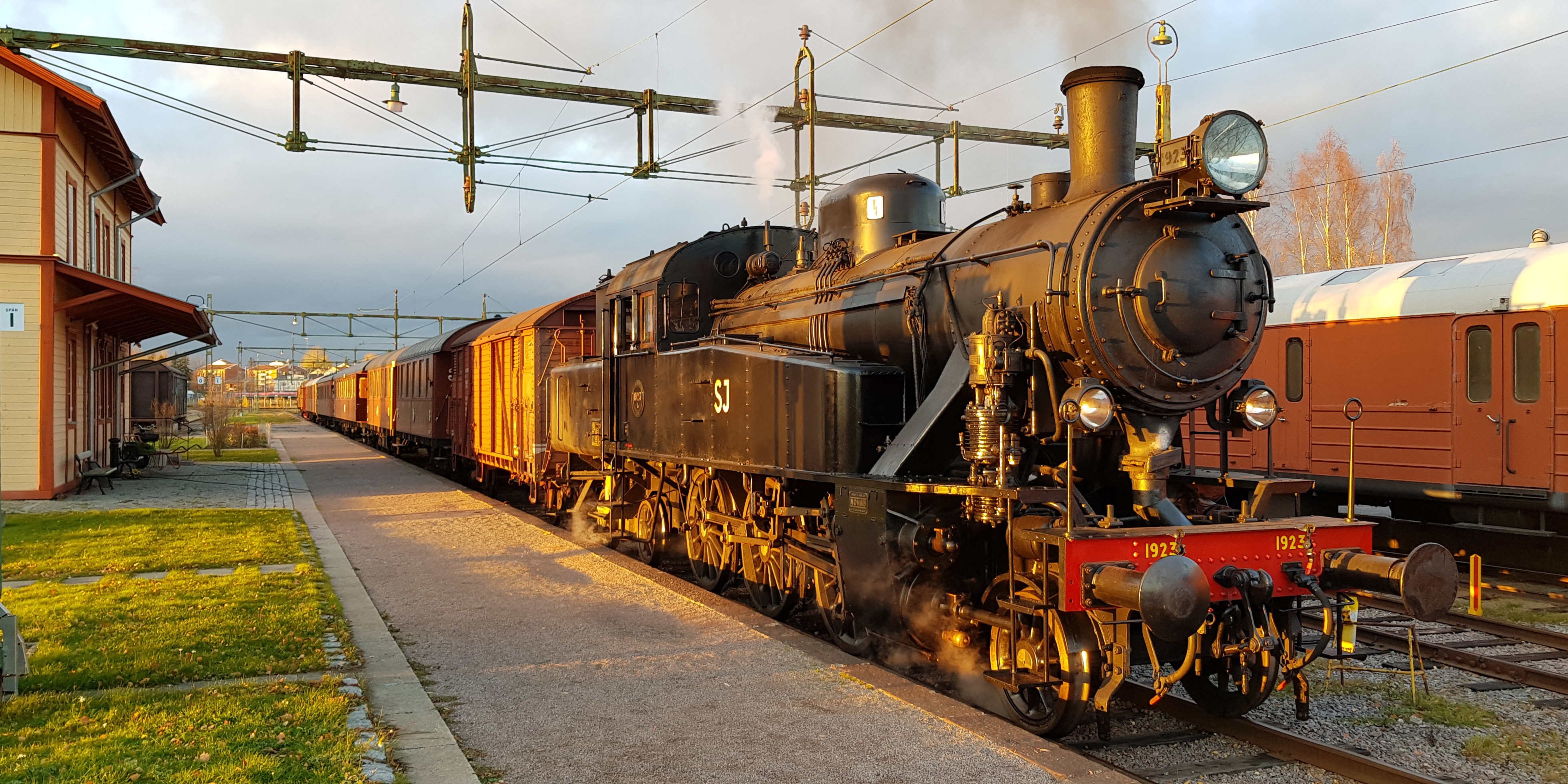 Lok ståendes vid perrong framför stationshus och puffar rök i kvällsljus.