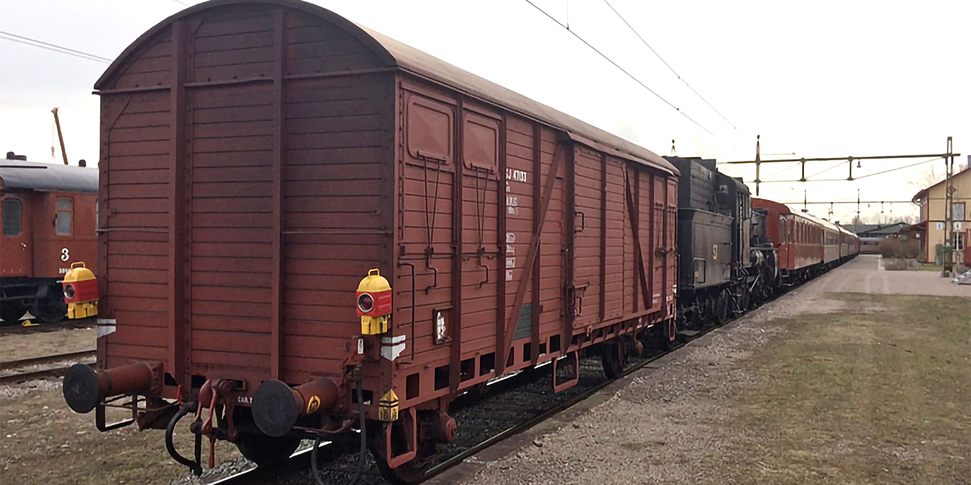 Bakändan på en rostbrun godsvagn med baklyktor som står sist i tåget vid en stationsbyggnad. 