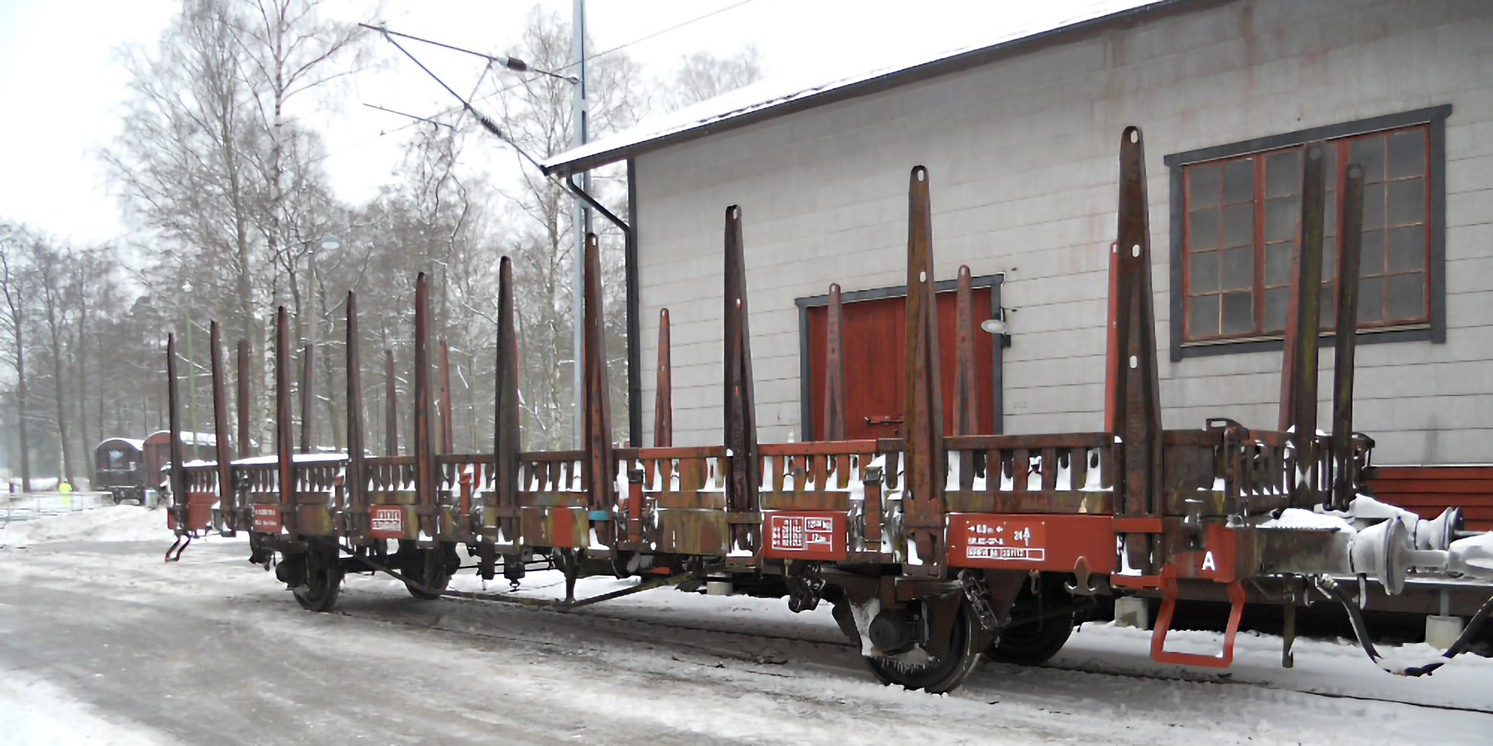 En öppen godsvagn med låga lämmar och stolpar står på ett spår framför en grå byggnad.
