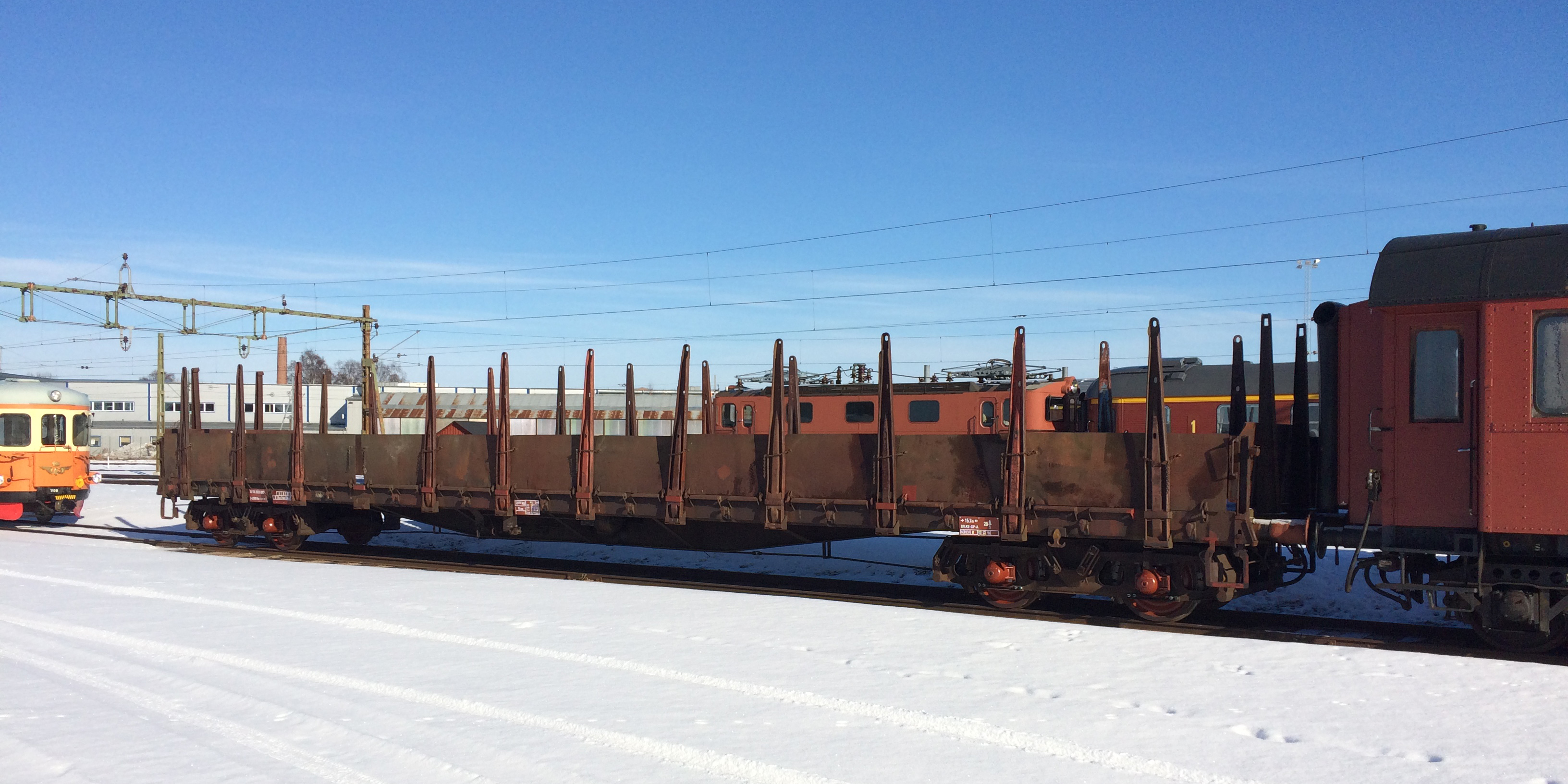 En låg öppen godsvagn som står på ett snötäckt spårområde omgiven av flera olika tåg.