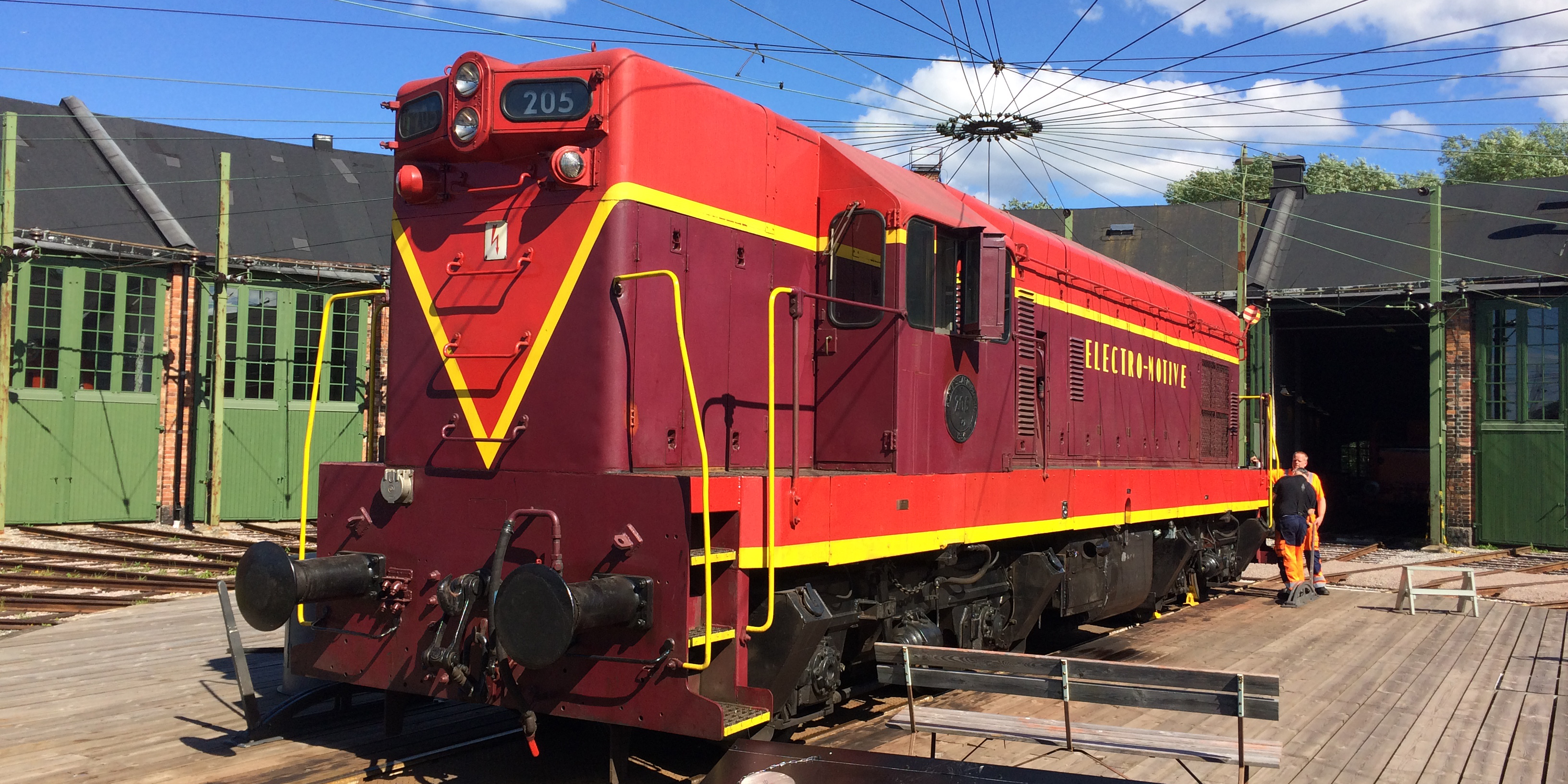 Large burgundy and red locomotive parked on a turntable outside the green round house. Yellow stripes on the locomotive.