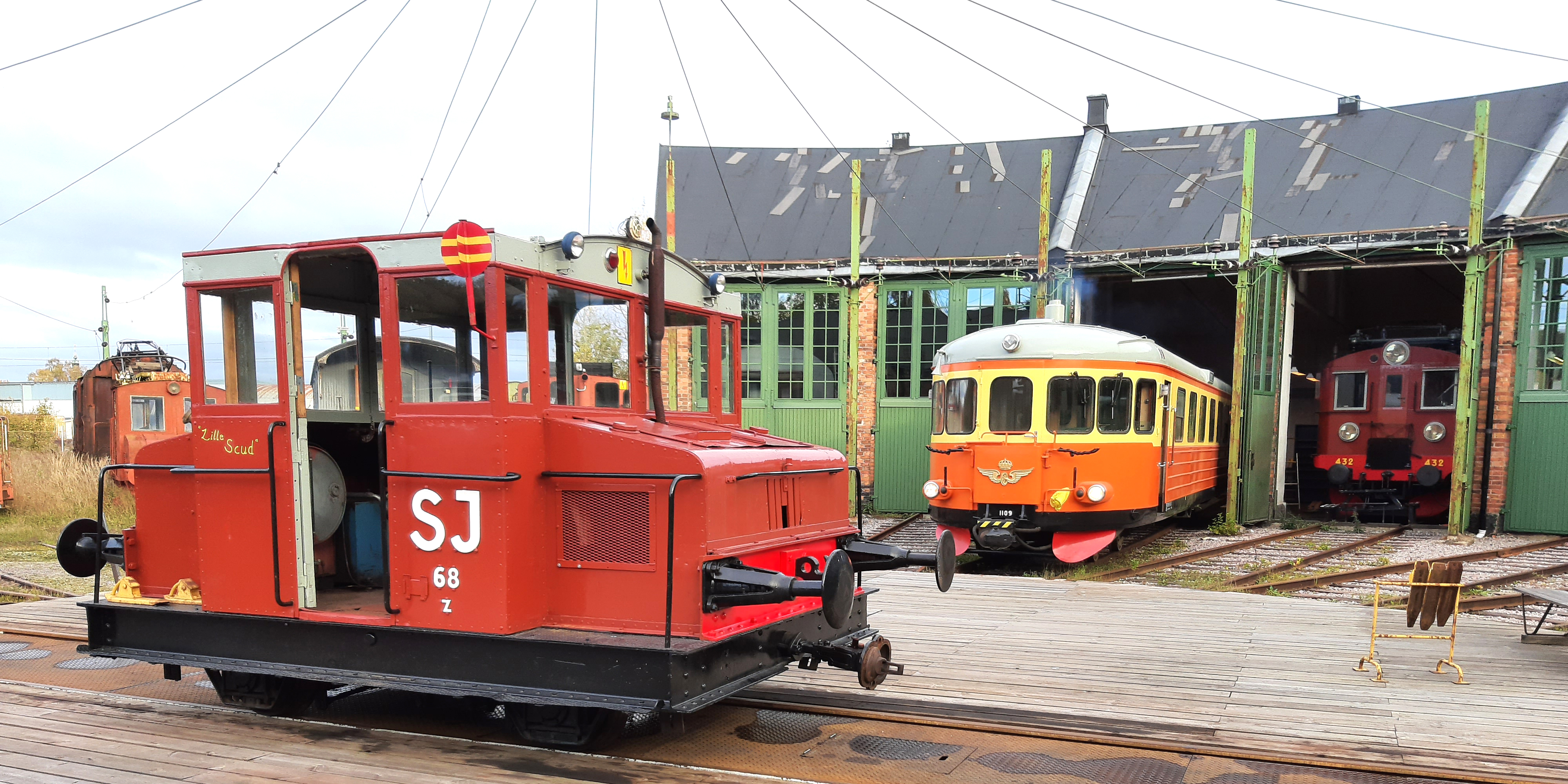 Rött lok framför grönt lokstall där rälsbuss tittar ut.