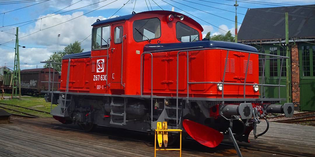 Rödmålat lok stående på räls framför grönt lokstall.