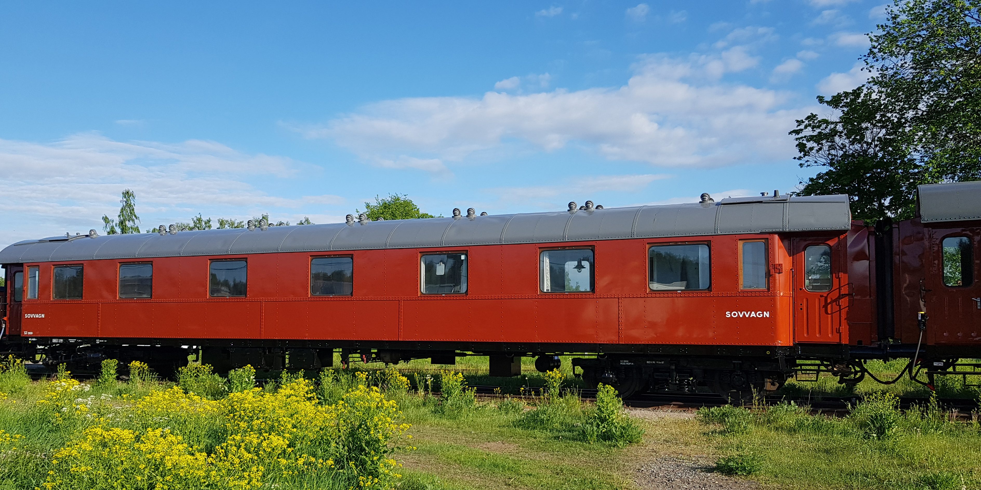 A red sleeping car on a rail lined with yellow meadow flowers over a clear blue sky.