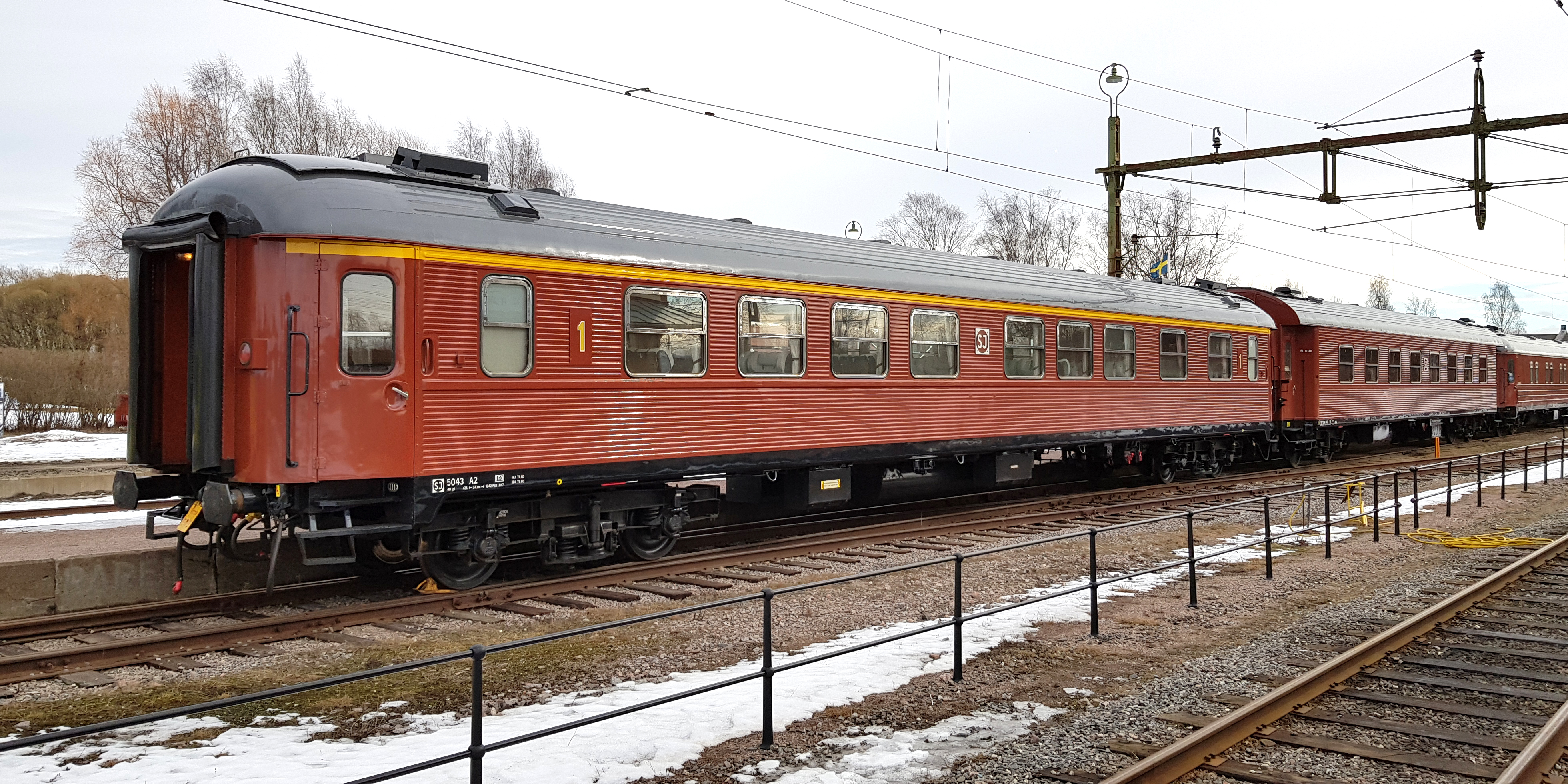 Roströd personvagn med gult streck ovanför fönstren kopplad sist i rad av vagnar.