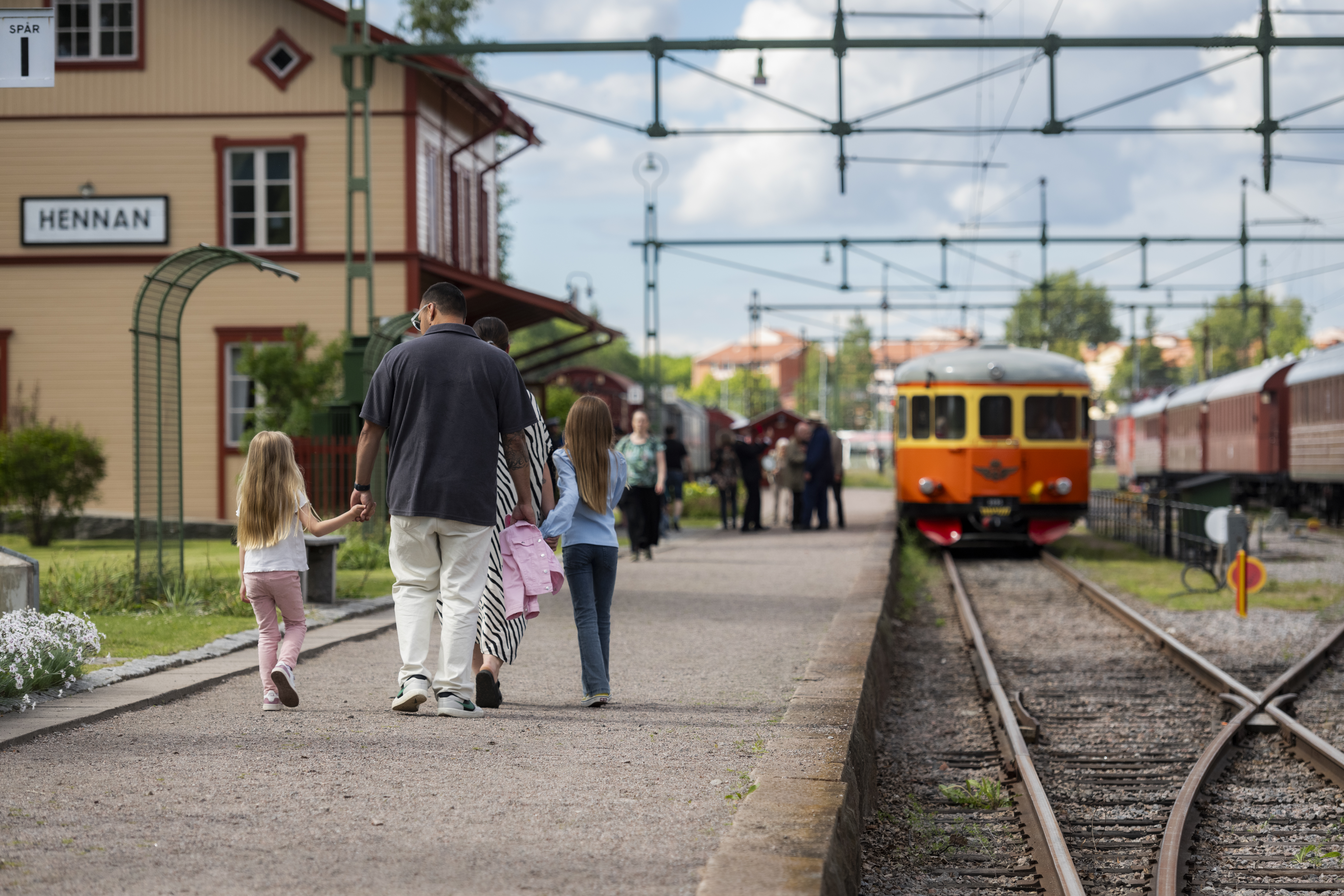 Perrong med grönska på ena sidan och en rälsbuss på andra. På perrongen går en pappa och håller en flicka i handen.