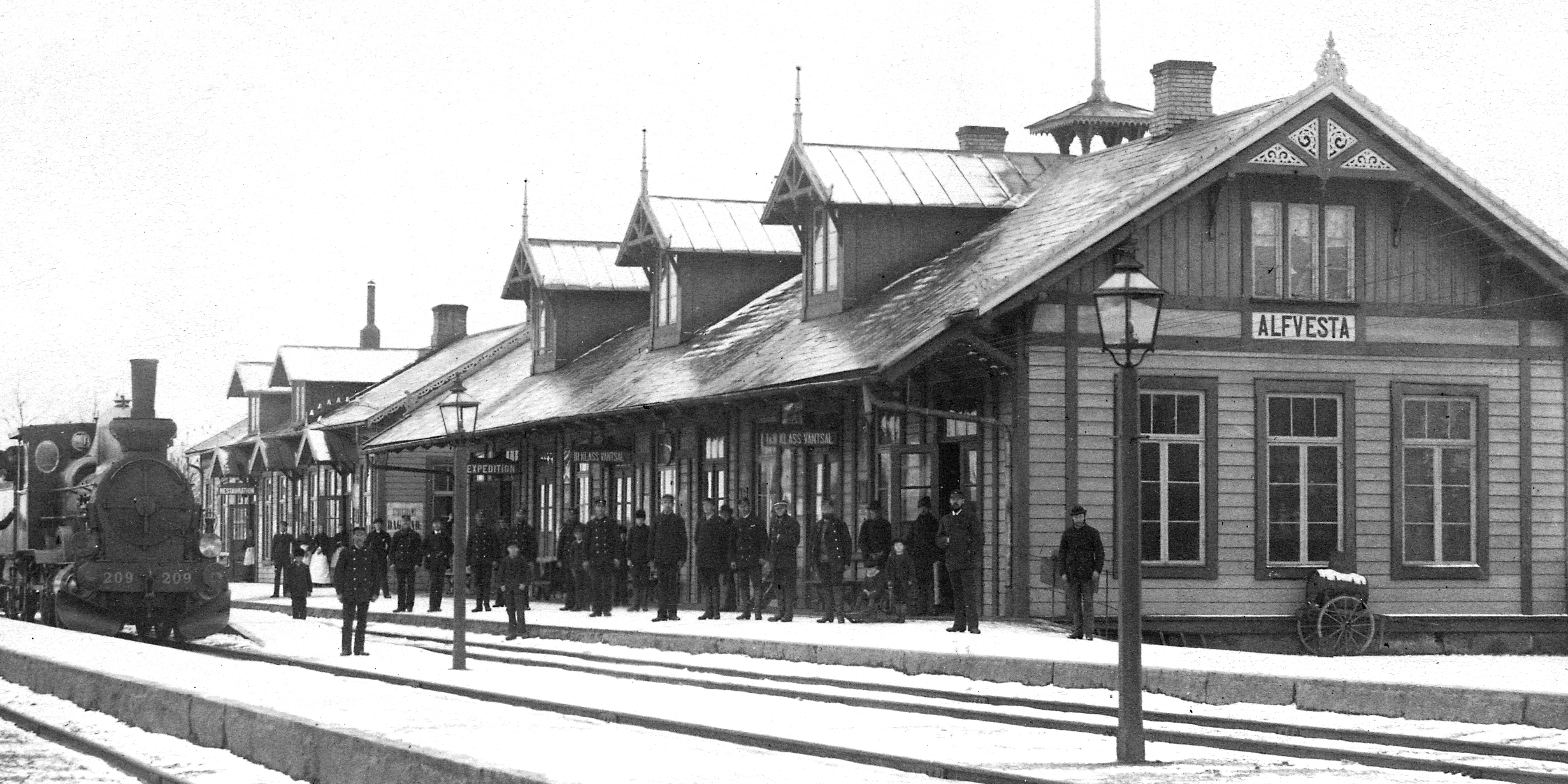 Många människor uppradade framför stort stationshus vid spår. Lok vid perrong.