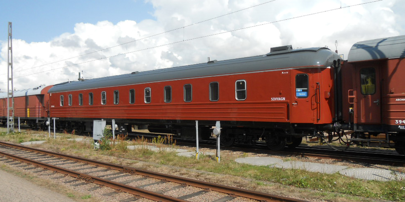 Red train with a black roof on the track with a sleeping car closest to it. An empty track in the foreground and green grass in between.