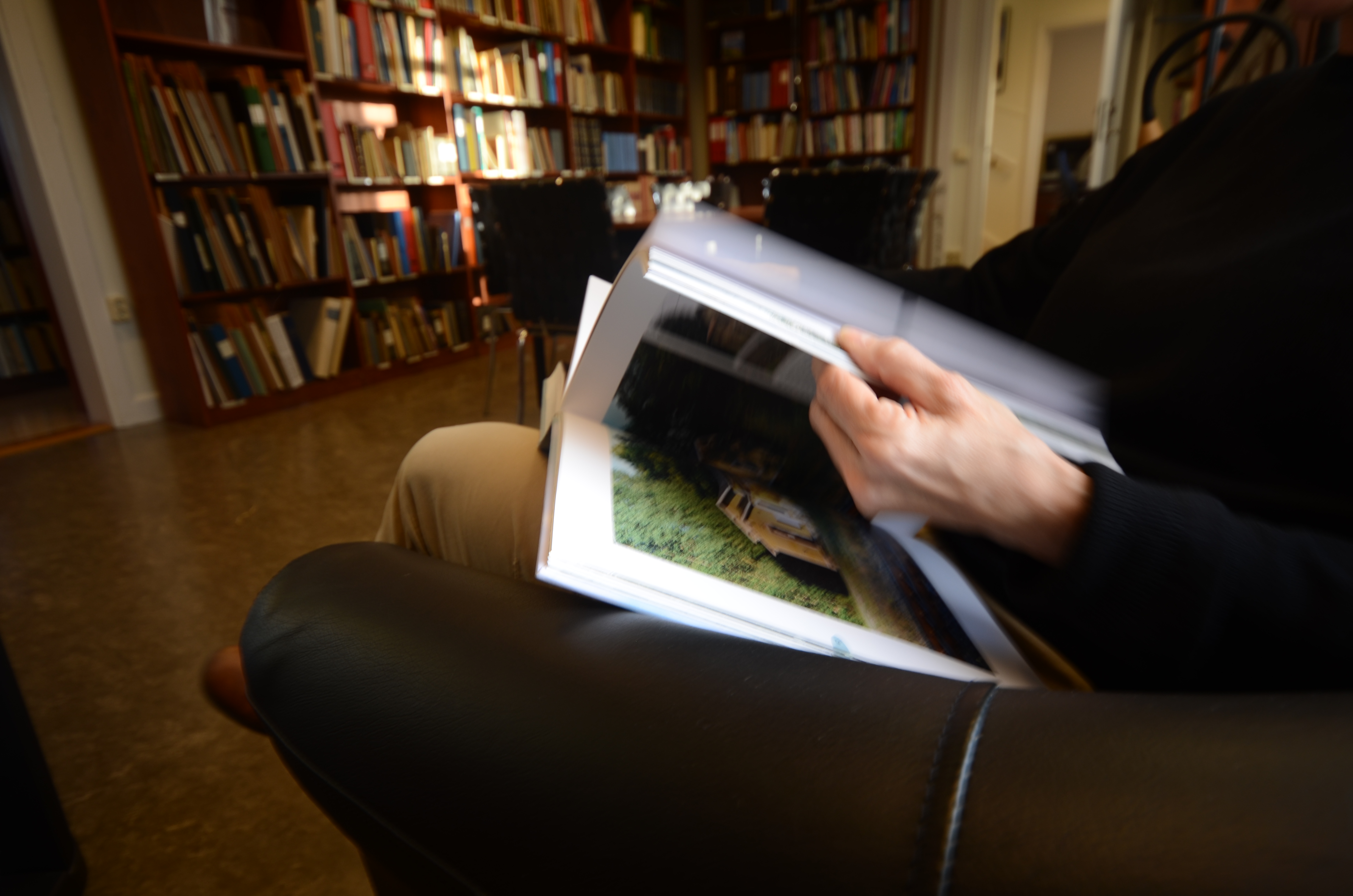 A person sitting in a leather armchair flips through a book with shelves of books in front of him.