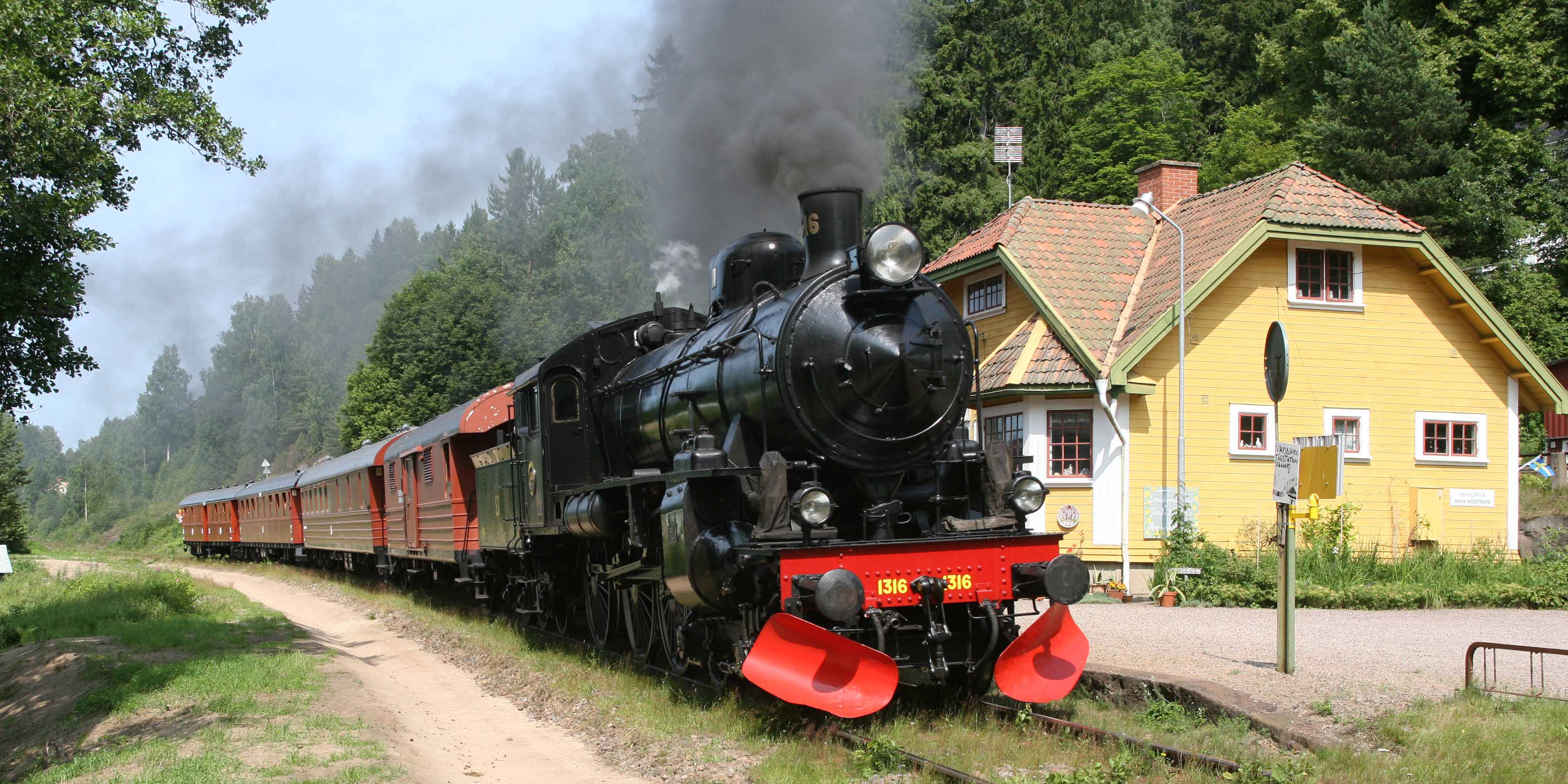 Black locomotive pulling fyra passenger cars on rails in front of yellow house. Puffs of black smoke.